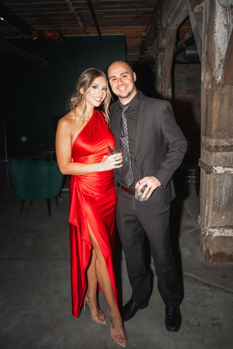 A woman in a red dress and a man in a black suit with a patterned tie pose together at an indoor event, both smiling and holding drinks.