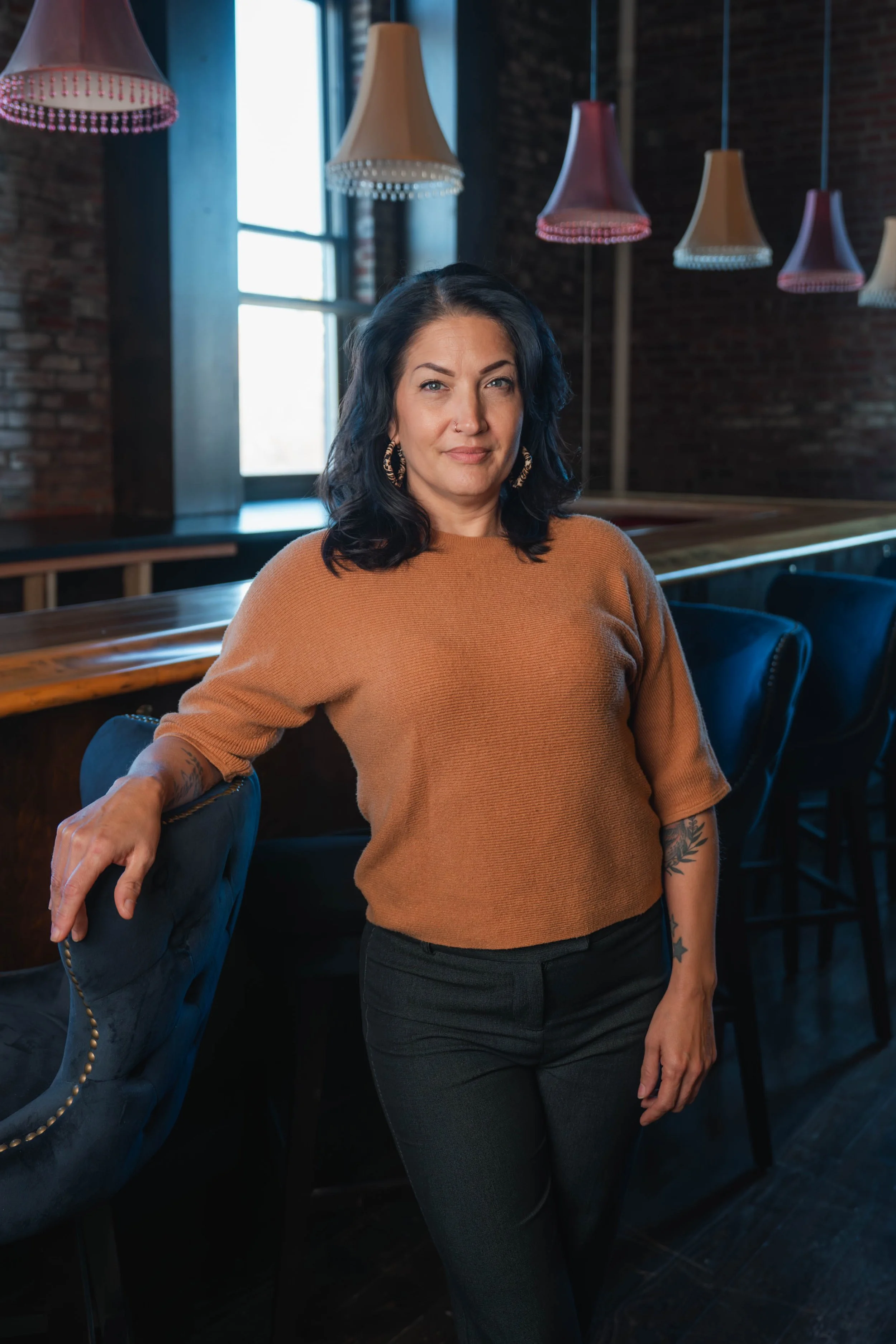 A woman with black wavy hair and hoop earrings posing in a dimly lit restaurant or bar with brick walls, large windows, and colorful pendant lights hanging from the ceiling.