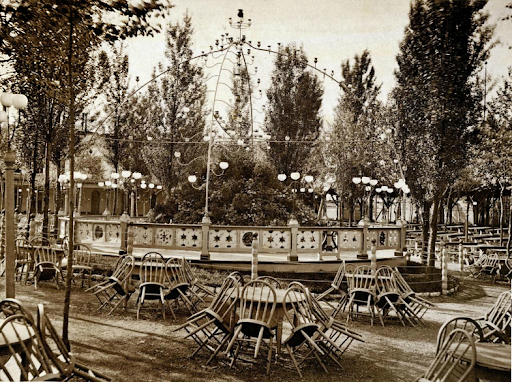 An old playground with a carousel in the center, surrounded by trees and empty chairs and tables, with string lights hanging overhead.