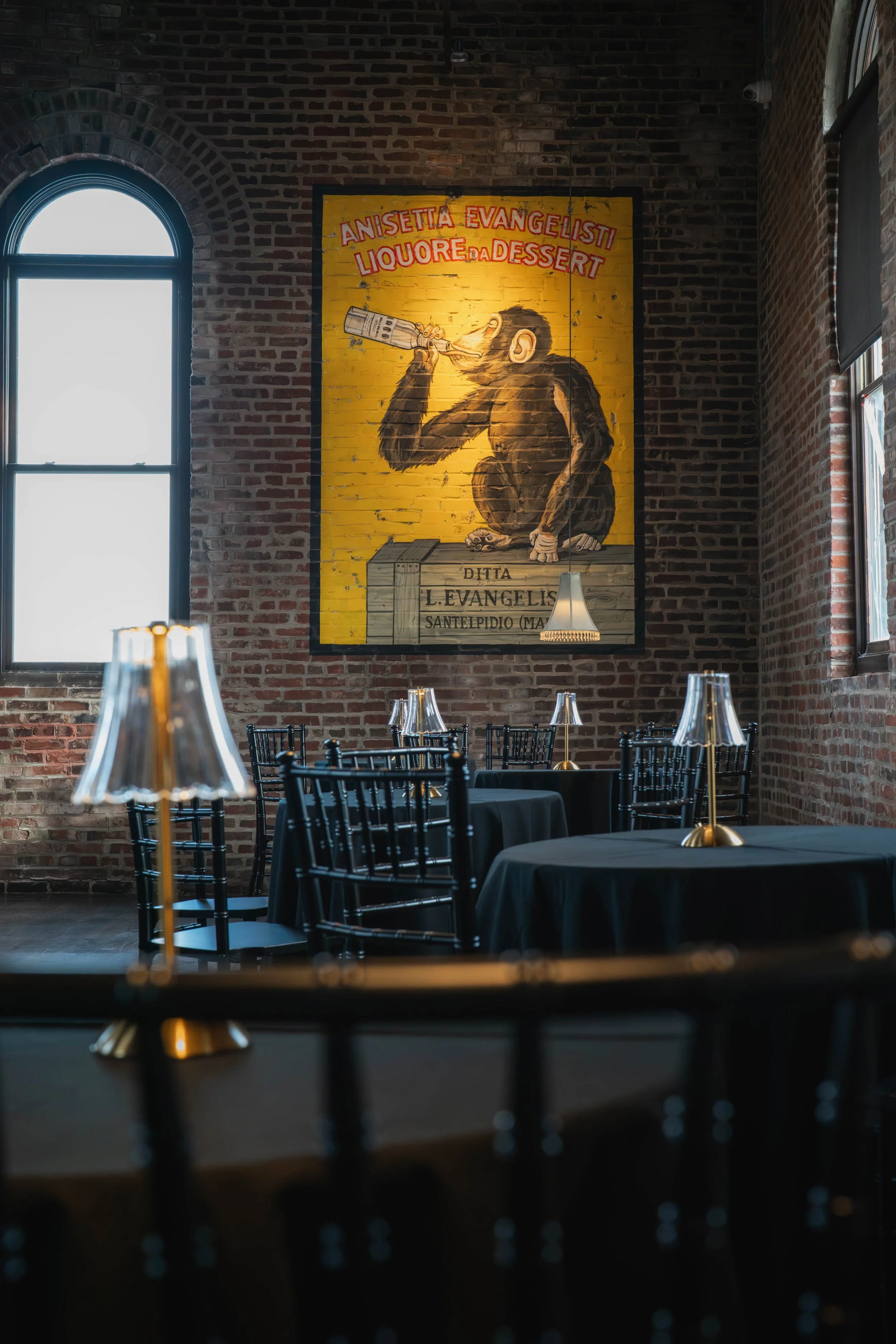 Interior of a restaurant with black tables, black chairs, and table lamps with transparent shades. A large, colorful mural of a monkey drinking from a bottle hangs on a brick wall. The restaurant has high windows and industrial-style lighting.