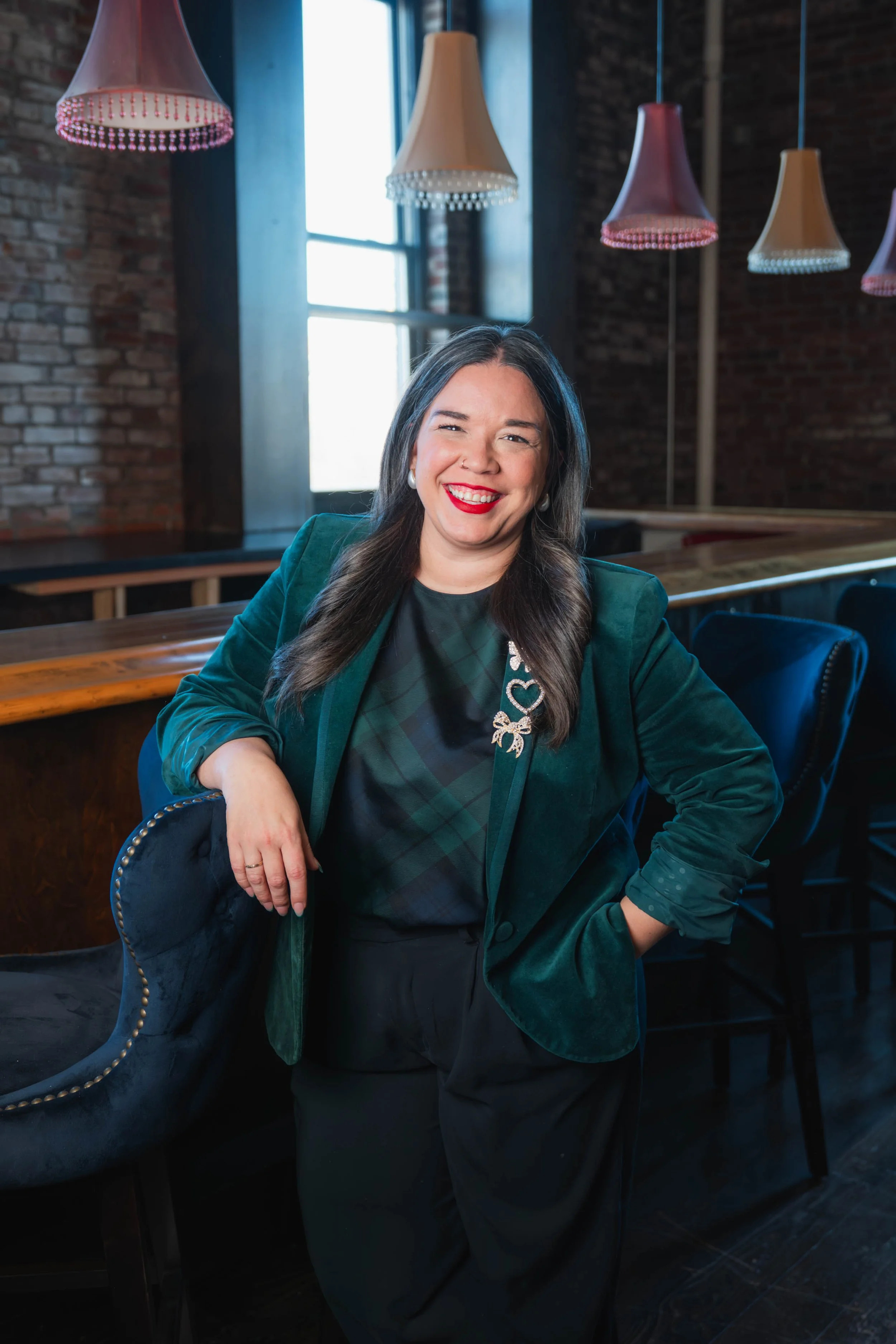 A woman with long dark hair wearing a green blazer, black top with a green plaid pattern, and black pants, smiling with red lipstick in a restaurant or bar with exposed brick walls, hanging pink and white lamps, and a large window in the background.