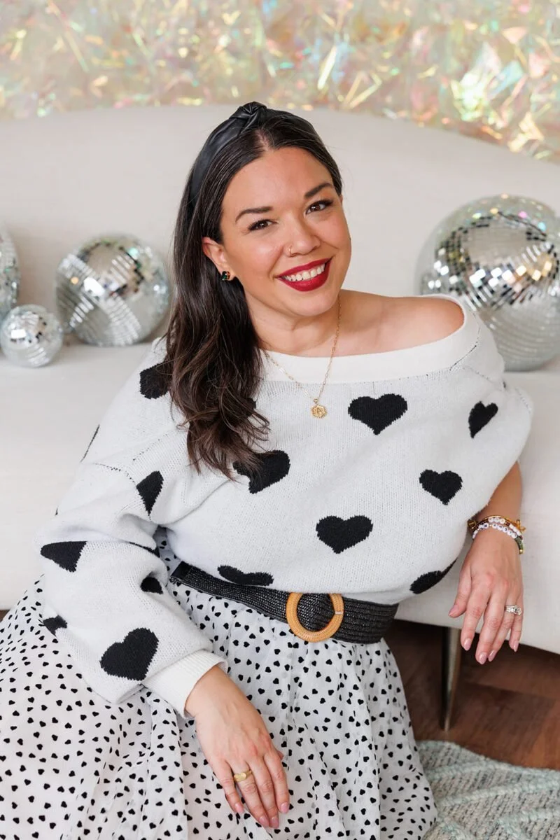 A woman smiling, sitting on a white couch with disco balls behind her, wearing a white sweater with black hearts, a polka dot skirt, and accessories.
