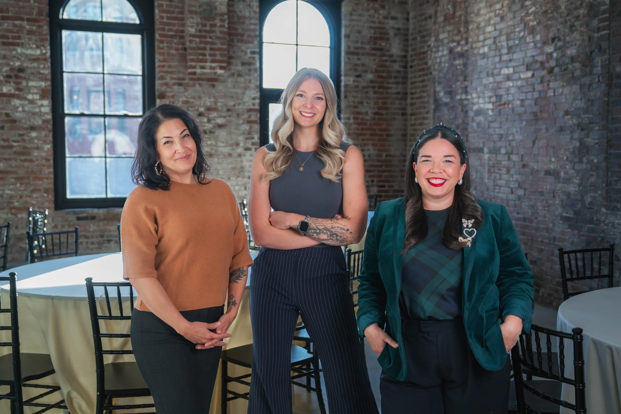 Three women standing in a room with brick walls and large windows, smiling at the camera.