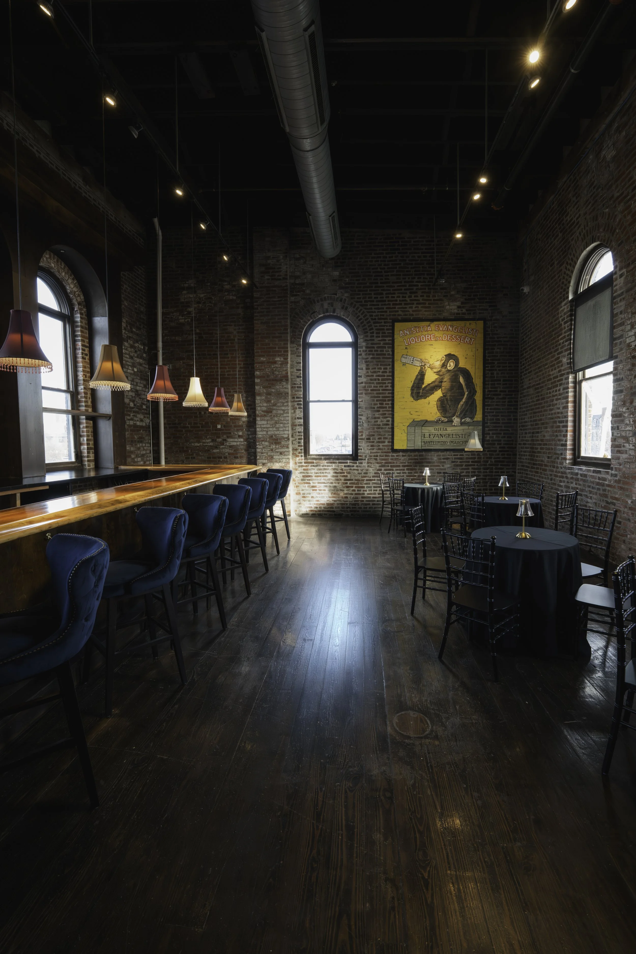 Interior of a restaurant or cafe with a brick wall, wooden floor, high windows, a bar with blue upholstered chairs, and round black tables with gold lamps. There is also a large colorful painting of a monkey drinking a beverage on the wall.