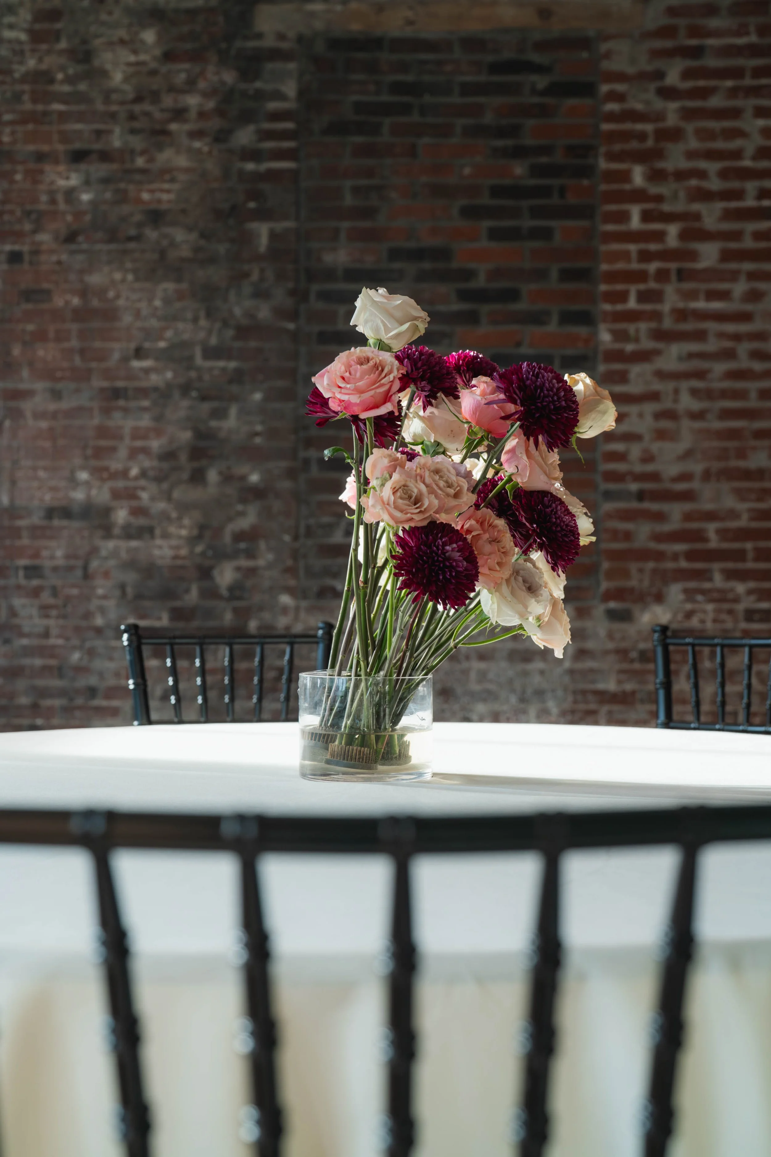 A glass vase with pink and white flowers and dark purple flowers on a white table in front of a brick wall, with black chairs around the table.