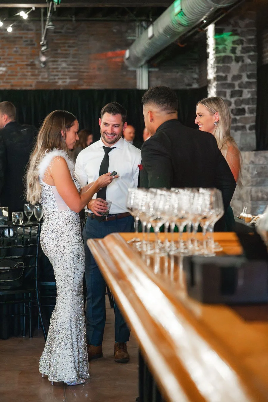 A group of four people dressed in formal attire, engaging in conversation at a social event, with wine glasses and a bar in the foreground, and exposed brick and industrial decor in the background.