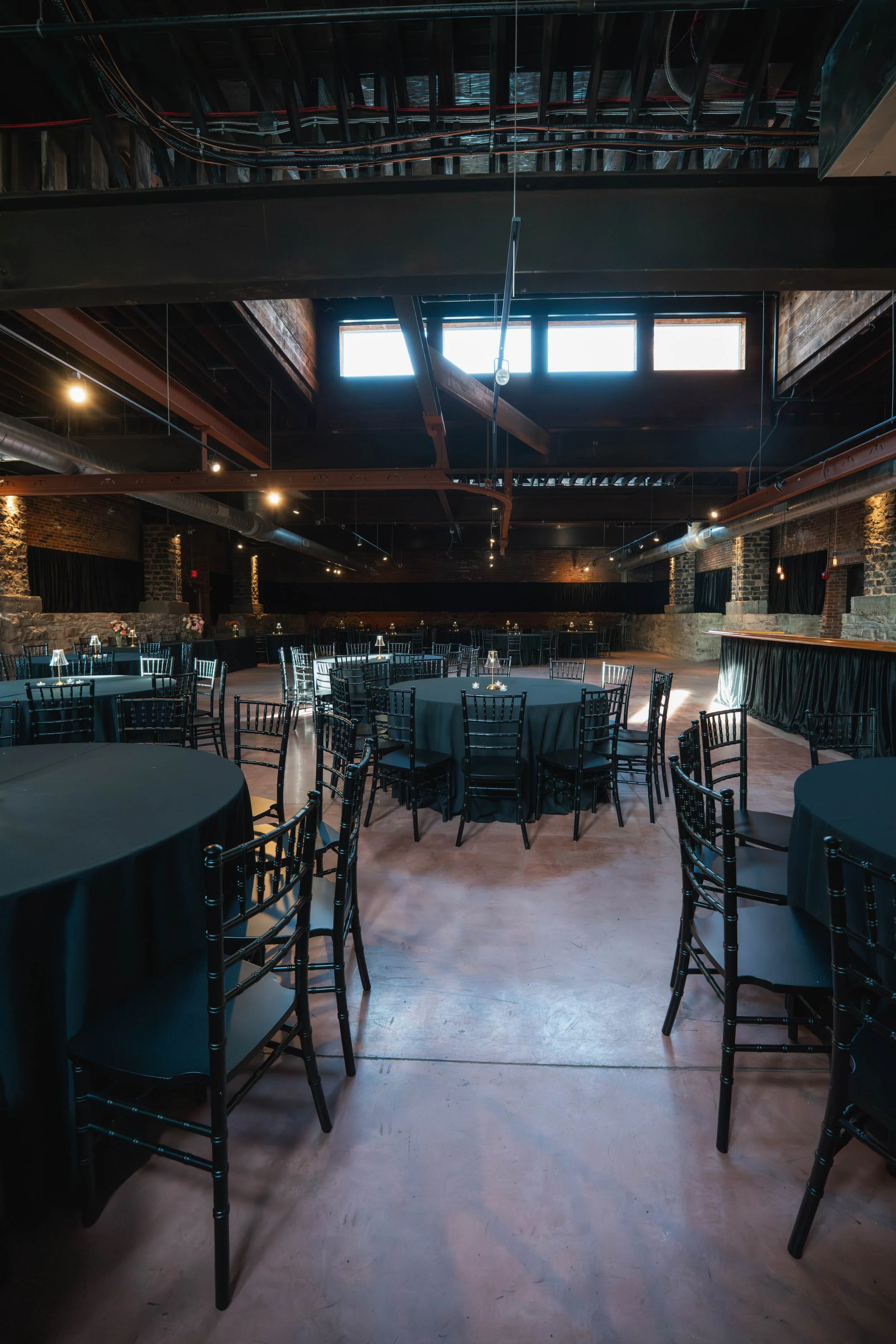 Empty event space with round tables covered in black tablecloths, black chairs, and small candle centerpieces, illuminated by natural light from windows and ceiling spotlights.