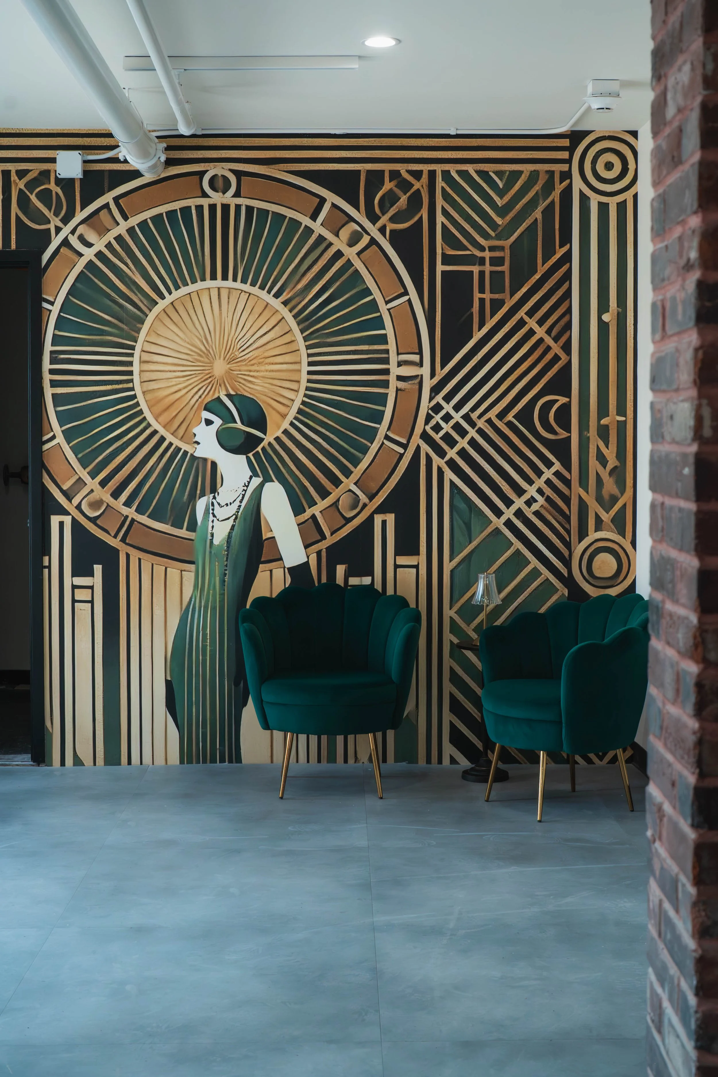 Interior view of a room with a large art mural of a vintage woman with a parasol in front of a clock, flanked by two green velvet armchairs, a small table with a lamp, and decorative geometric patterns.