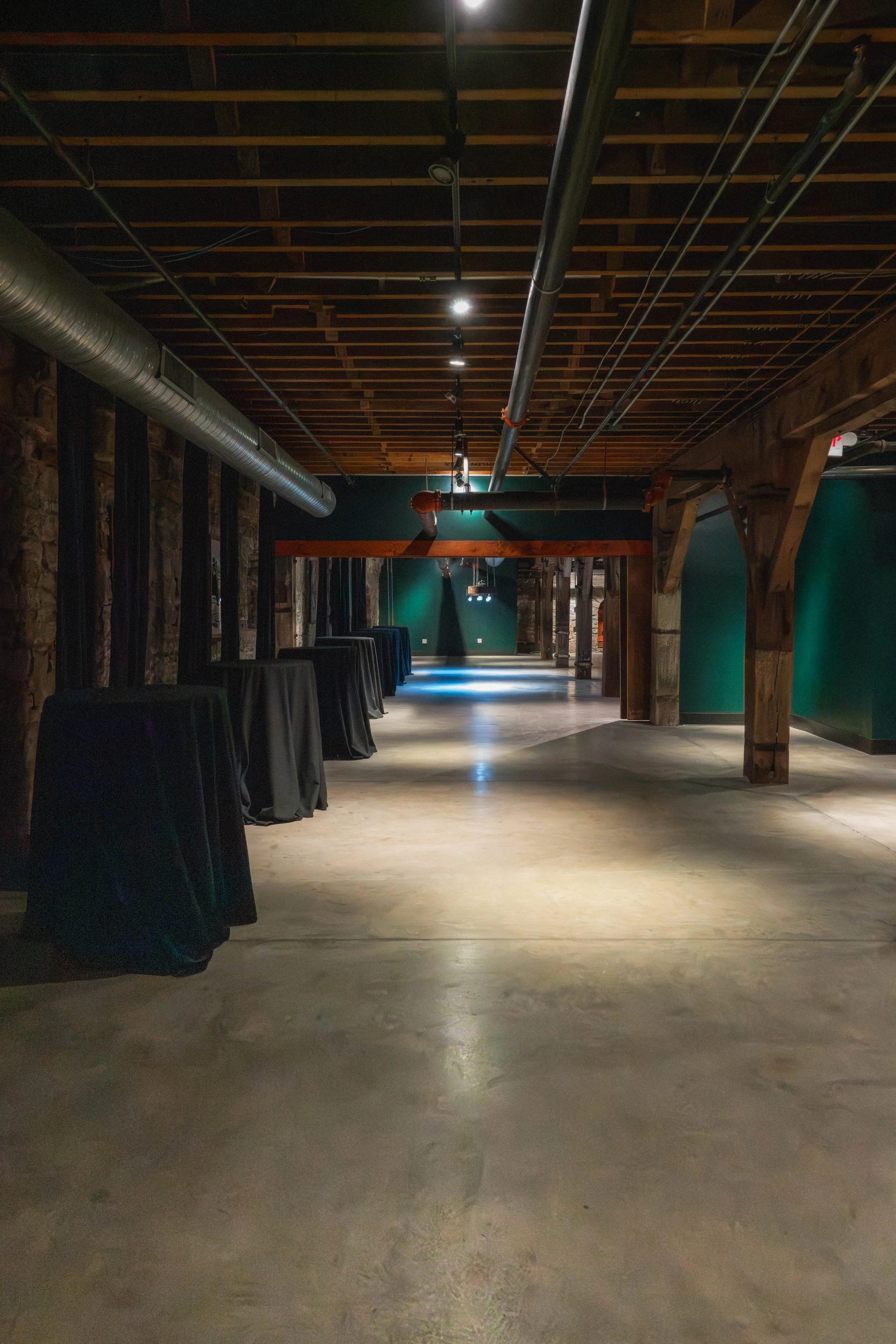 Empty event space or hall with high wooden ceiling, exposed pipes, and dark green and brick walls, with tables covered in black tablecloths lined along the side.