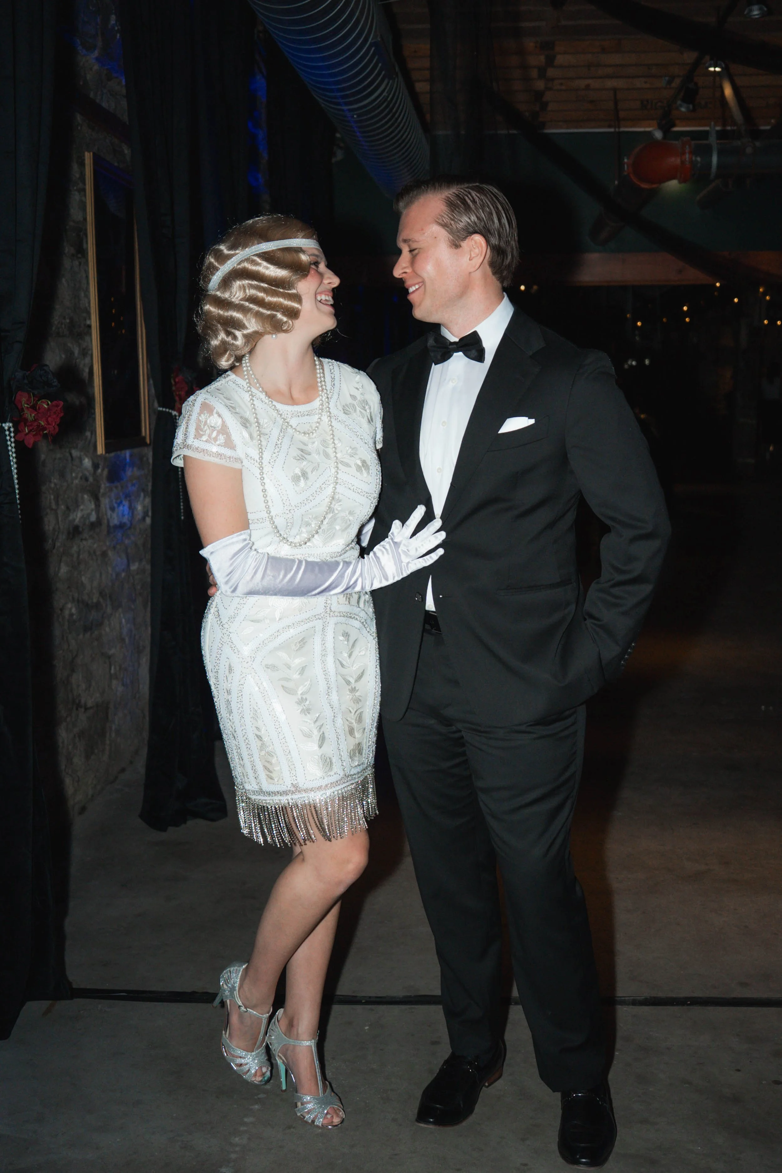 A couple dressed in 1920s style vintage clothing, smiling and looking at each other at a themed event or party.