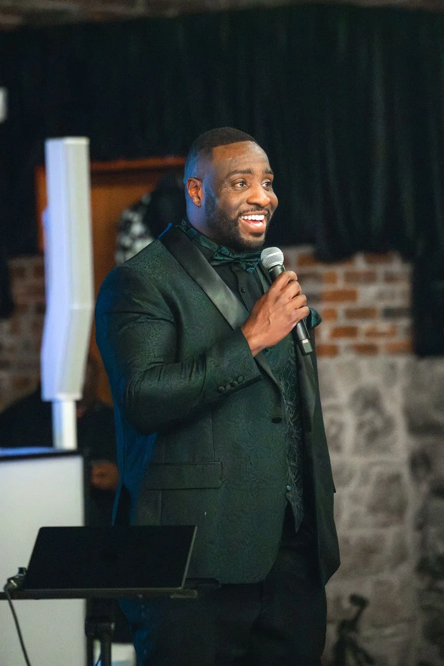A man in a black tuxedo with a satin lapel and bow tie, smiling and speaking into a microphone at an indoor event with a brick wall and dark curtains in the background.