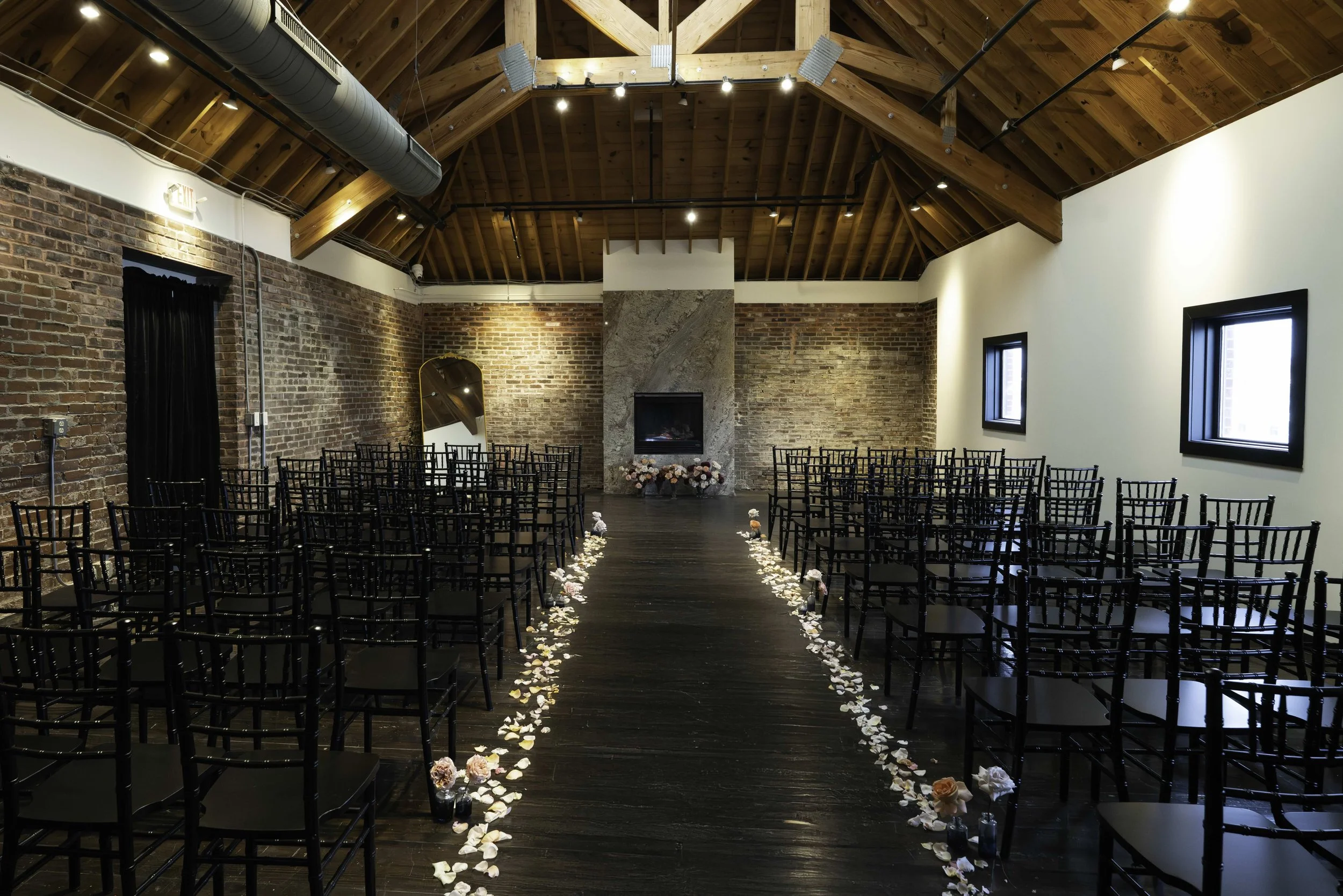 Indoor wedding ceremony setup in a modern rustic style with black chairs arranged along an aisle decorated with rose petals and flowers, leading to a fireplace at the front of the room with exposed brick walls and wooden vaulted ceiling.