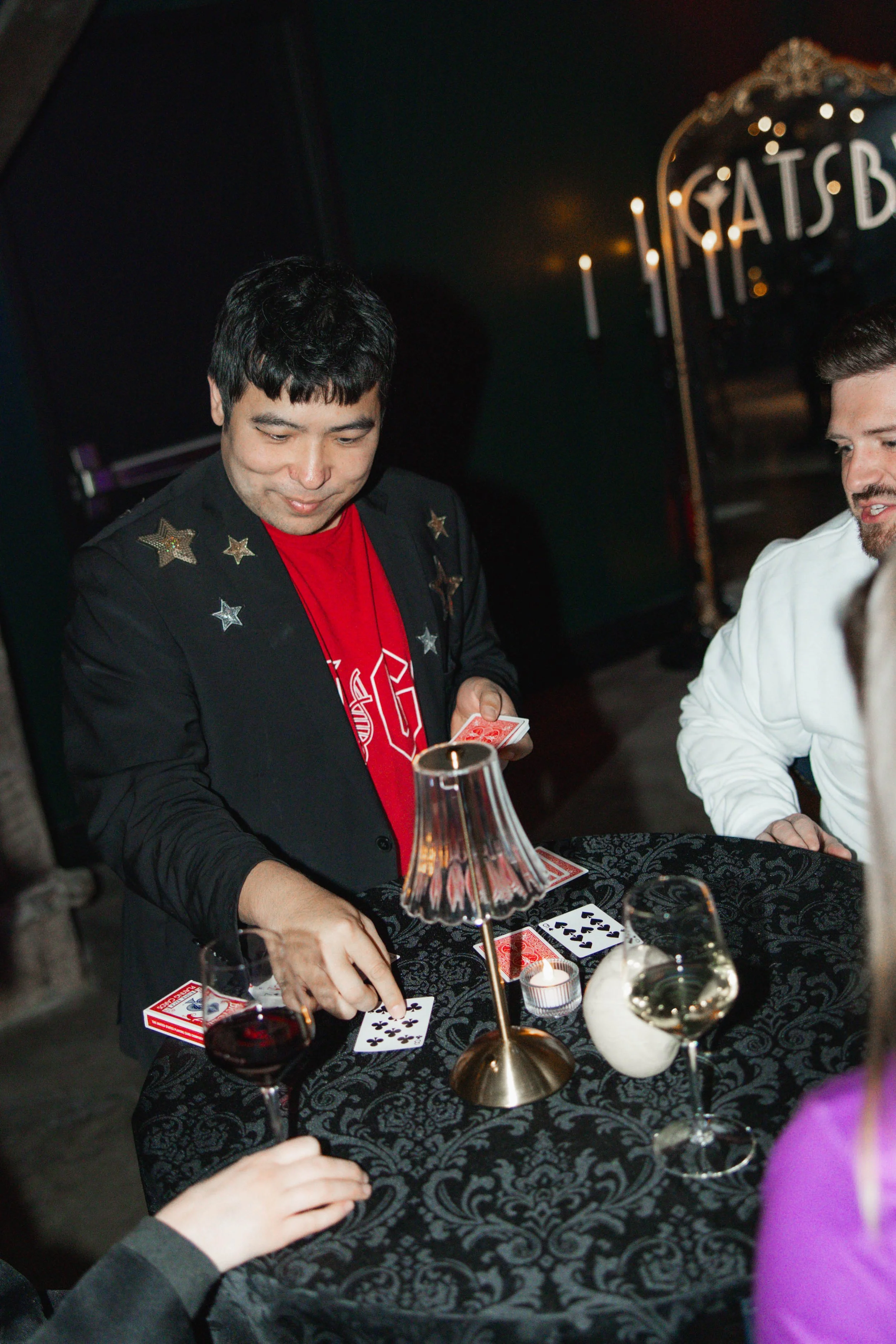 A man in a black jacket with star patches playing cards with others at a table with a black tablecloth, a small lamp, and wine glasses.