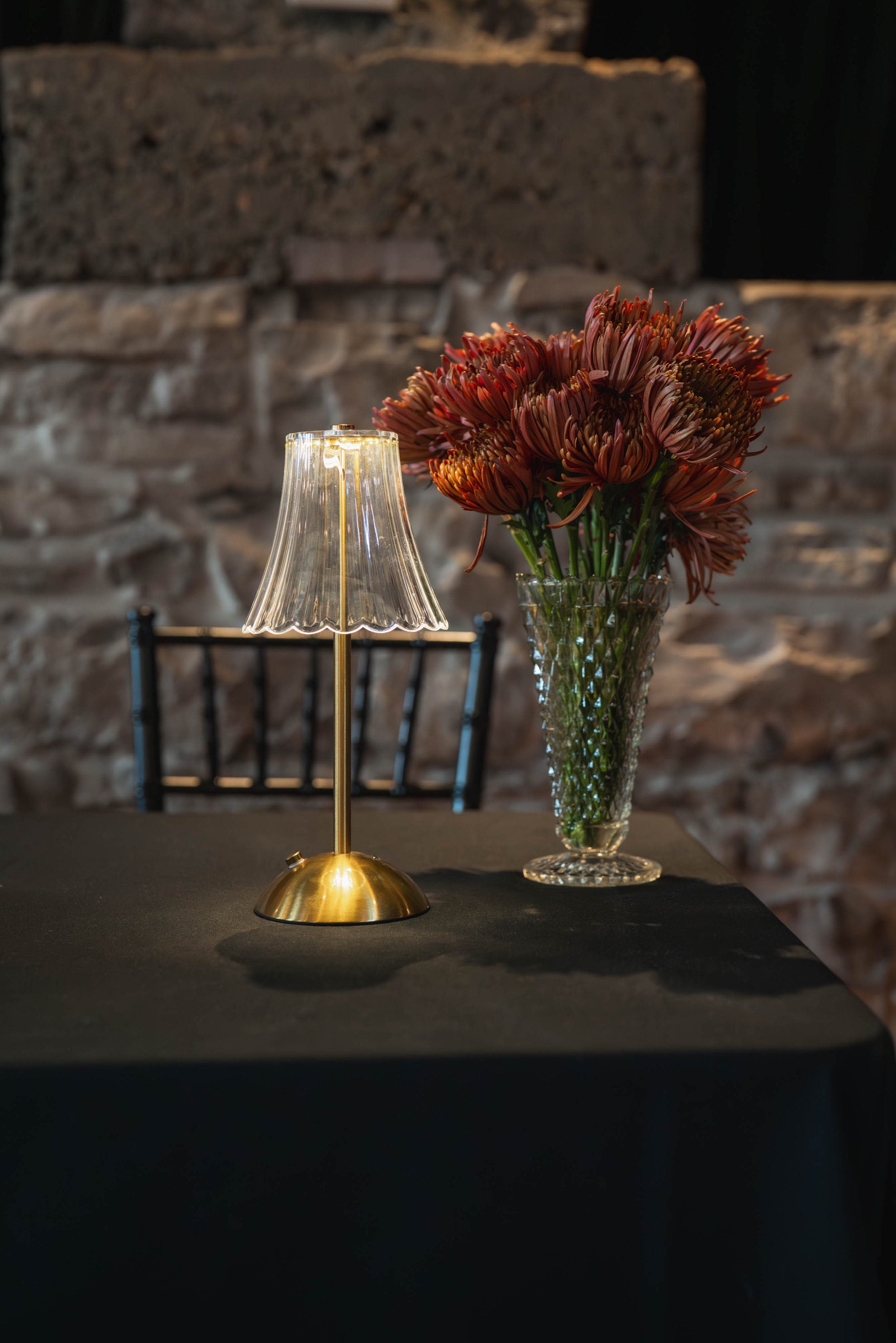 A small lamp with a clear, ribbed shade and a brass base, placed on a table next to a tall glass vase filled with pink and red flowers, with a stone wall background.