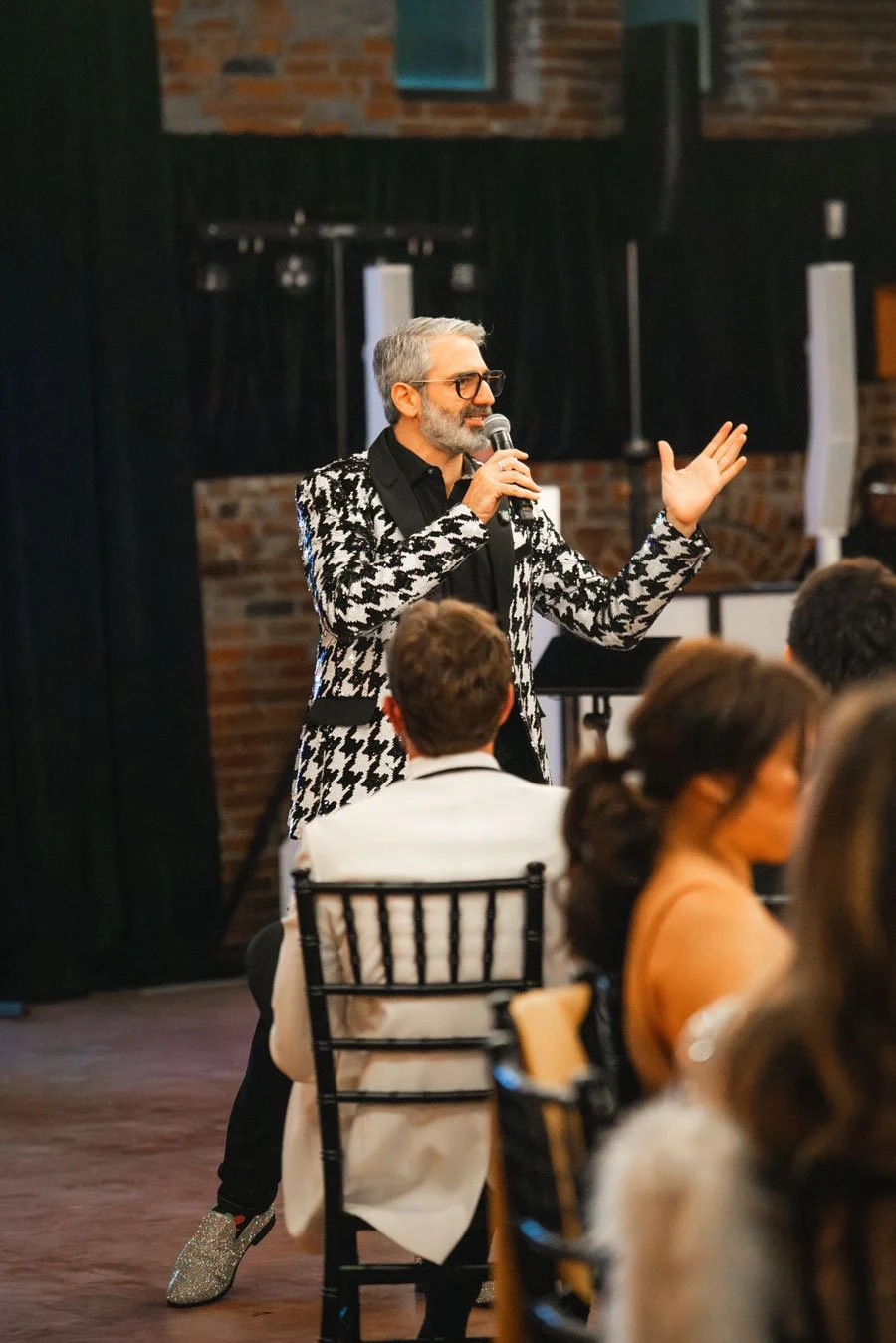 A man with gray hair, glasses, a beard, wearing a black and white houndstooth blazer, speaking into a microphone at an indoor event with seated attendees.