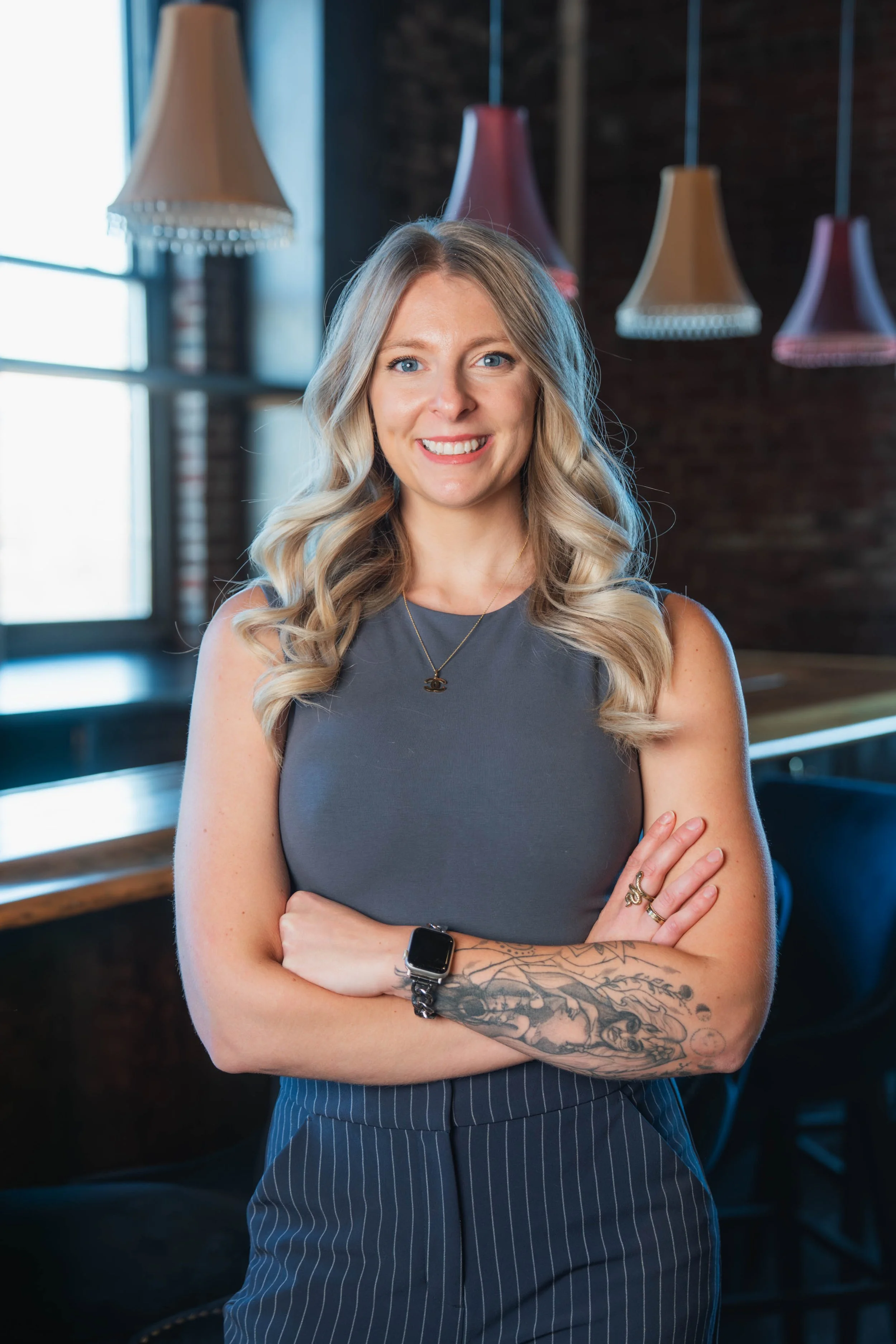A woman with blonde curly hair, wearing a sleeveless gray top, standing with crossed arms in a modern indoor setting with hanging lamps and large window.
