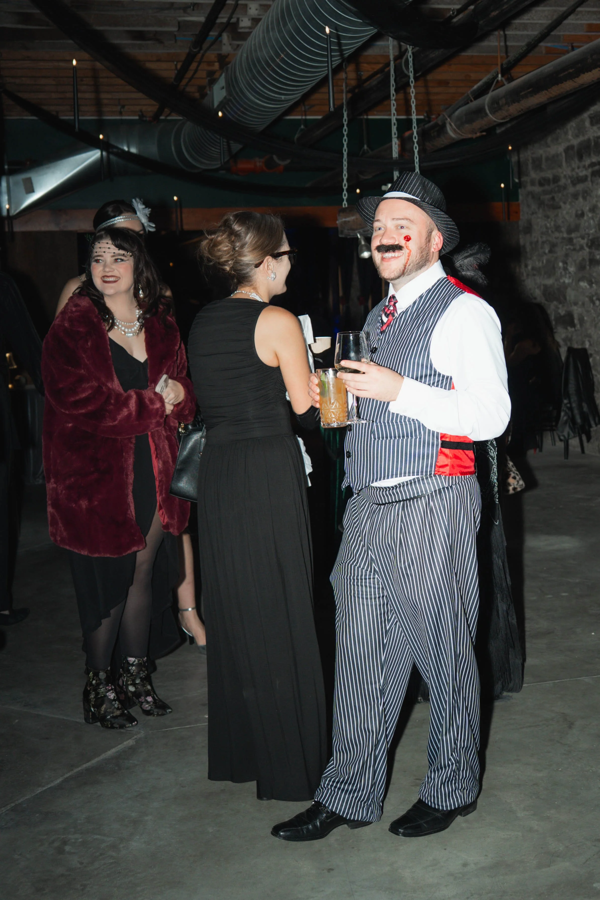 Group of people dressed in vintage 1920s costumes at a themed party, with a man in a pinstripe suit and fedora, woman in flapper dress, and others in vintage attire, holding drinks in an industrial-style venue.