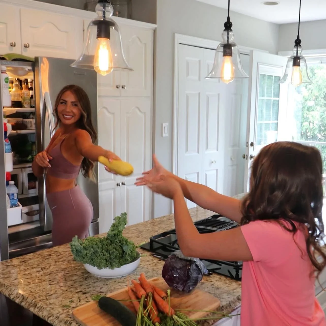 A woman in athletic wear reaching out to receive a yellow sponge from a young girl in a pink shirt in a bright kitchen.