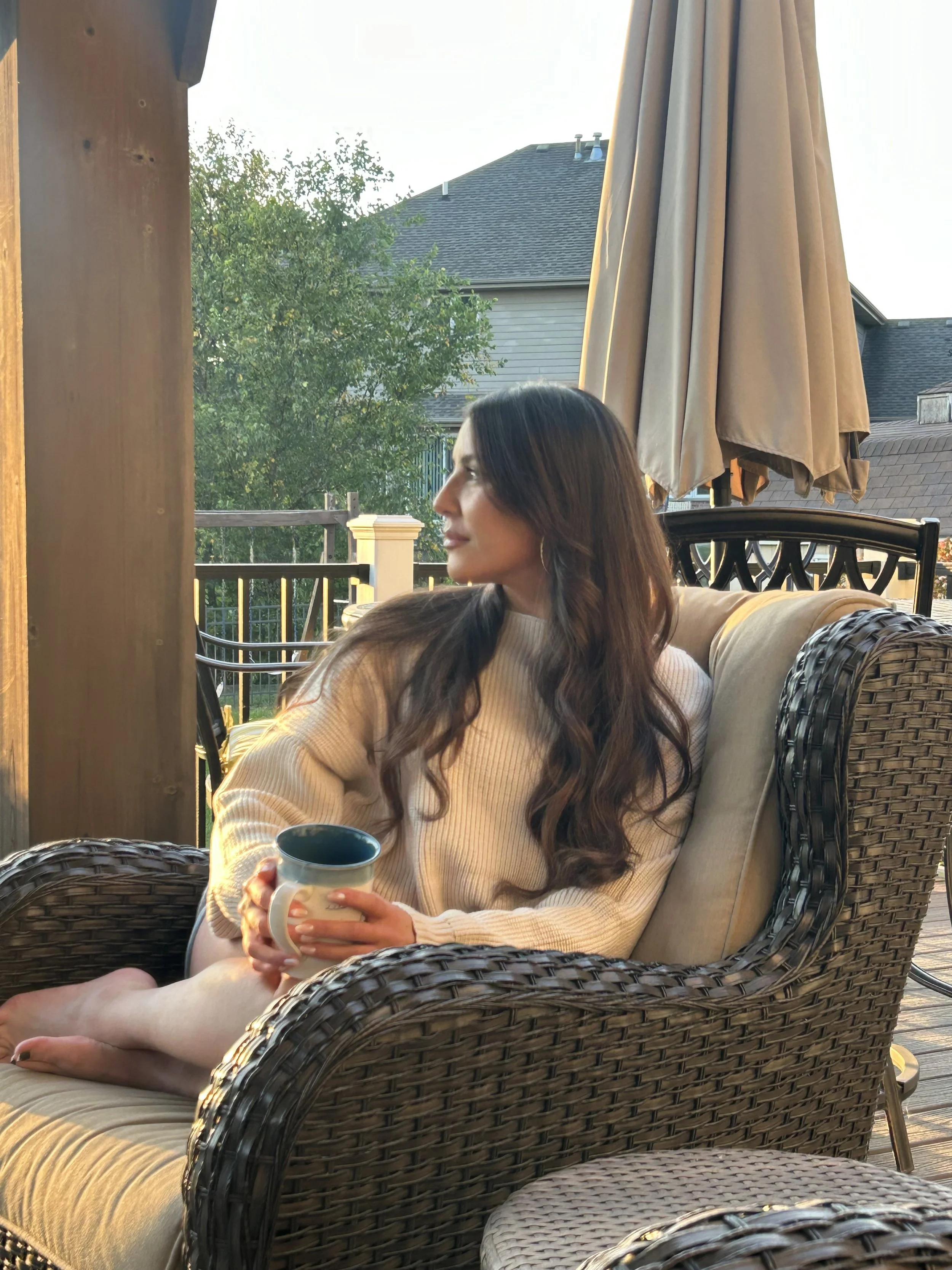 A woman with long dark wavy hair, wearing a cream-colored sweater, sits on an outdoor wicker sofa holding a mug, looking to her right. The scene is set on a wooden deck with a large umbrella, a house with a dark roof, and trees in the background during the daytime.