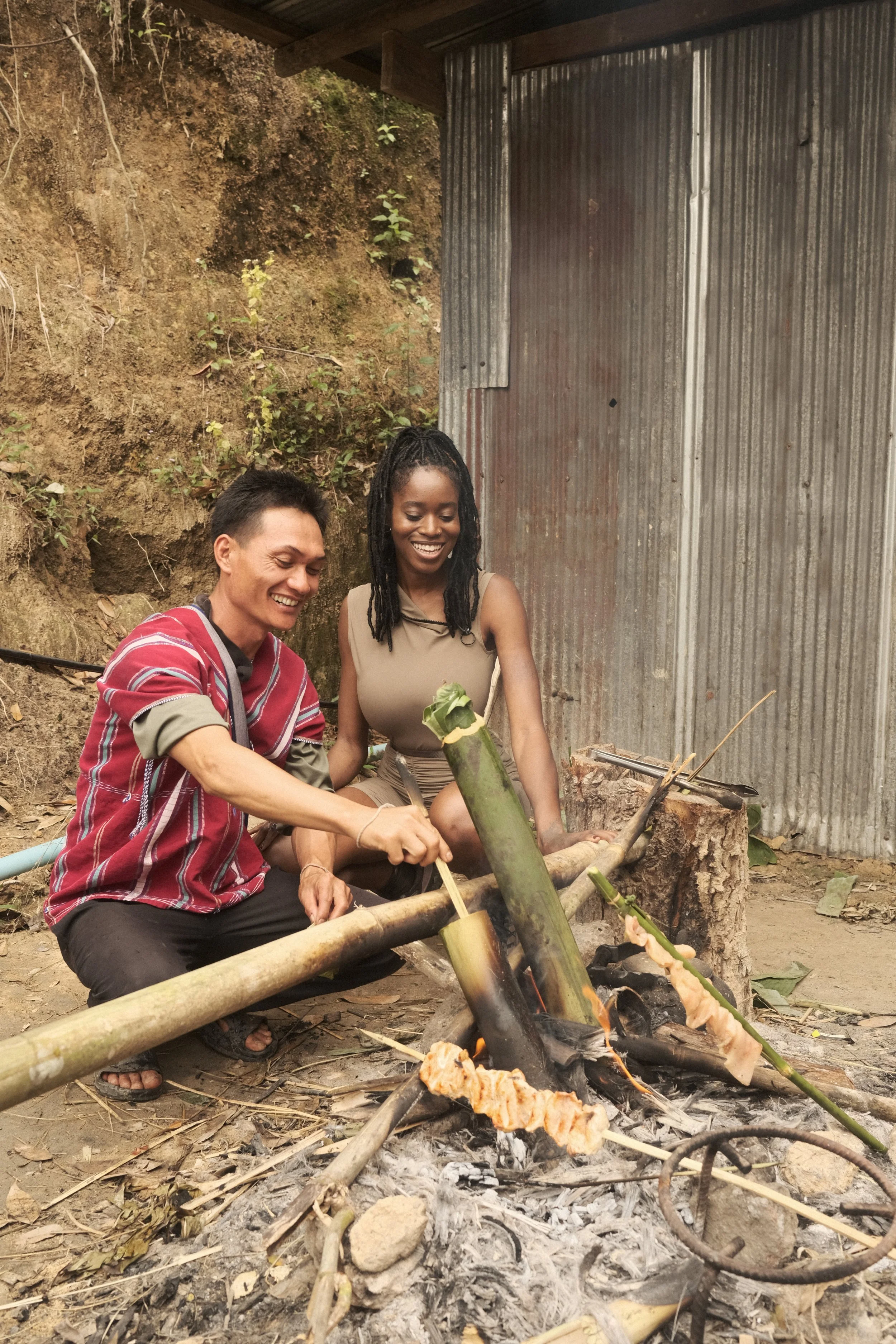Two people are roasting food over an open fire outdoors, with a rustic backdrop of a dirt ground, a hillside, and a corrugated metal wall. They appear happy and engaged.