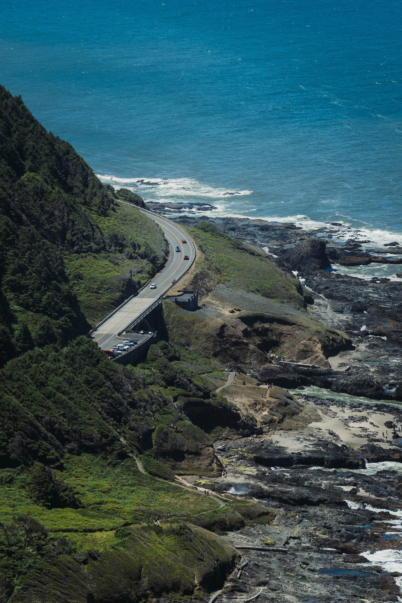 Coastal highway winding along a steep hillside overlooking the ocean with rocky shores and waves.
