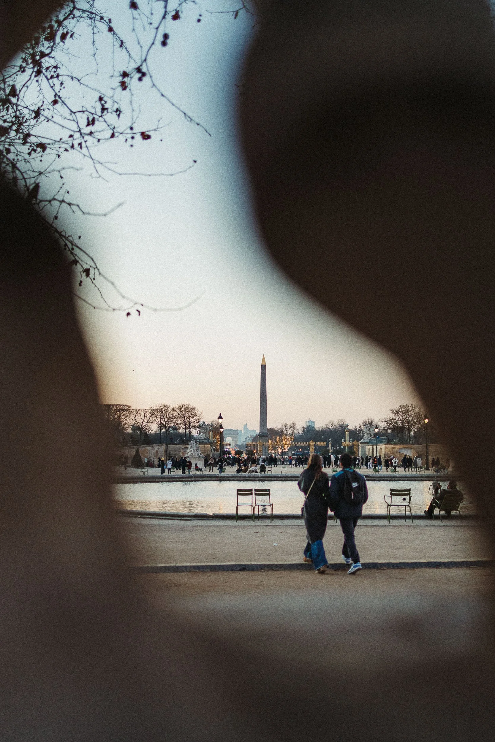 View of the Washington Monument in a park, seen through a frame created by two blurred dark objects. People are walking and sitting near a body of water, with trees and a cityscape in the background.