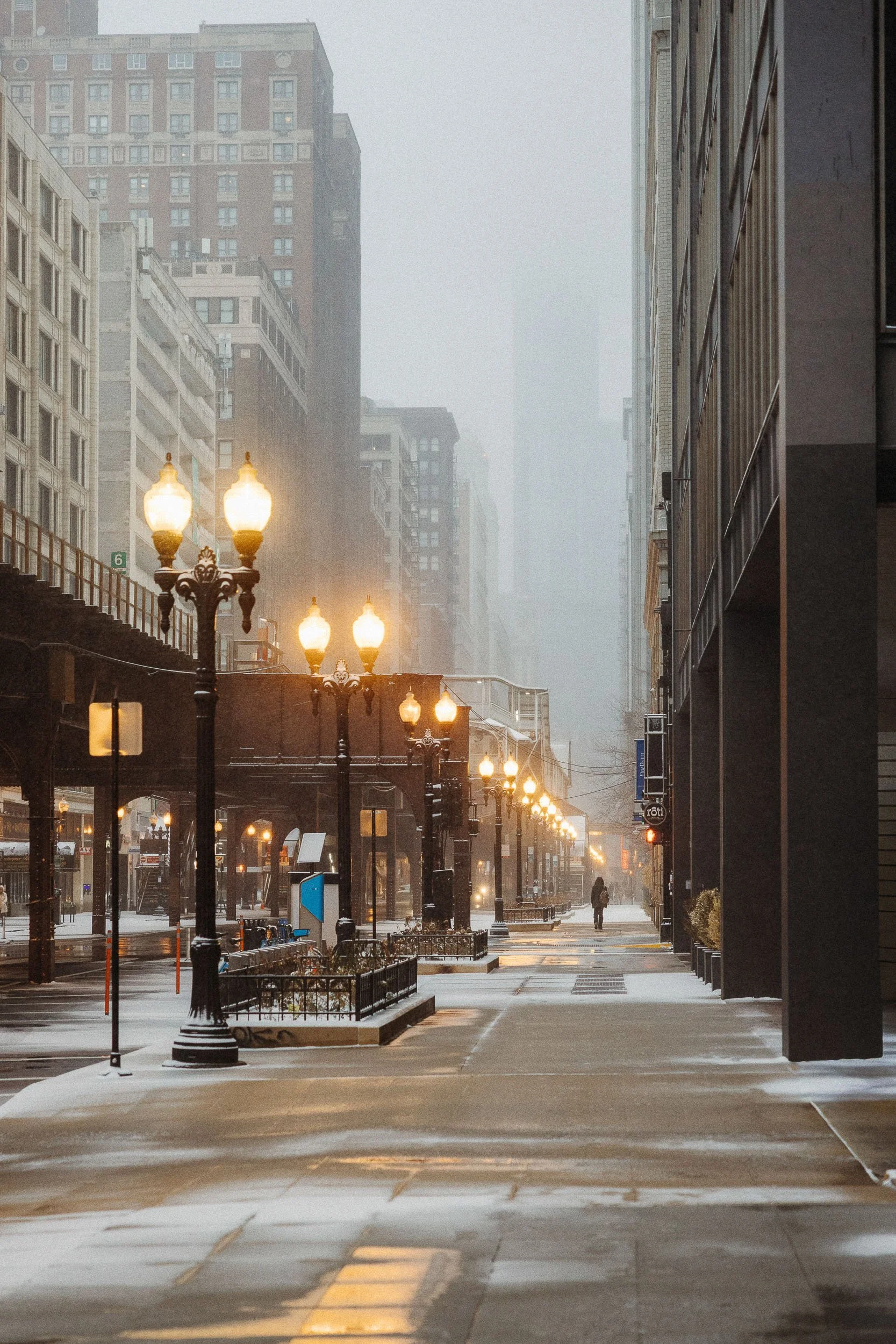 Empty city street with lit lampposts, snow on the ground, tall buildings, and foggy atmosphere.
