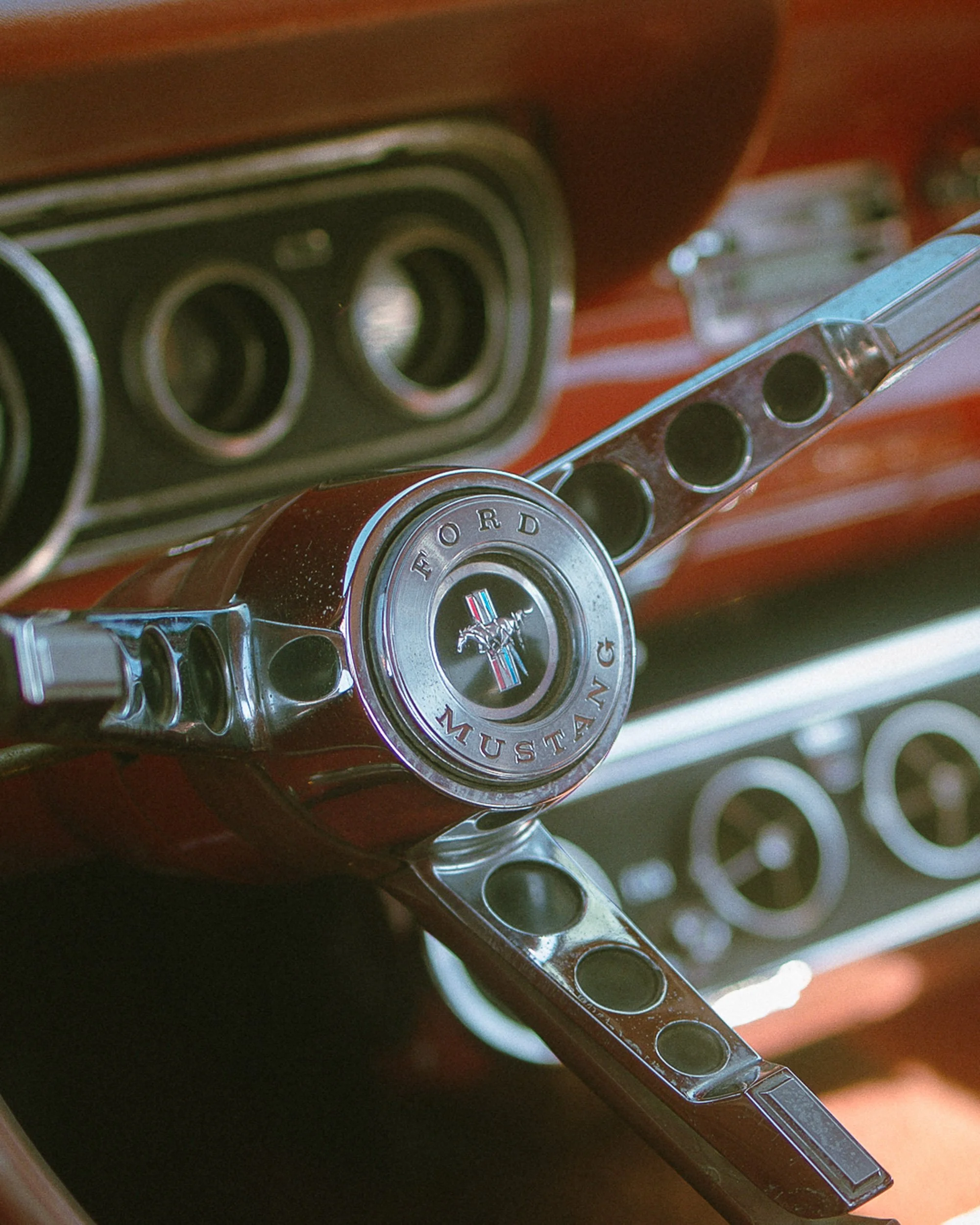 Close-up of a vintage Ford Mustang steering wheel with chrome spokes and Mustang emblem in the center, with dashboard gauges in the background.
