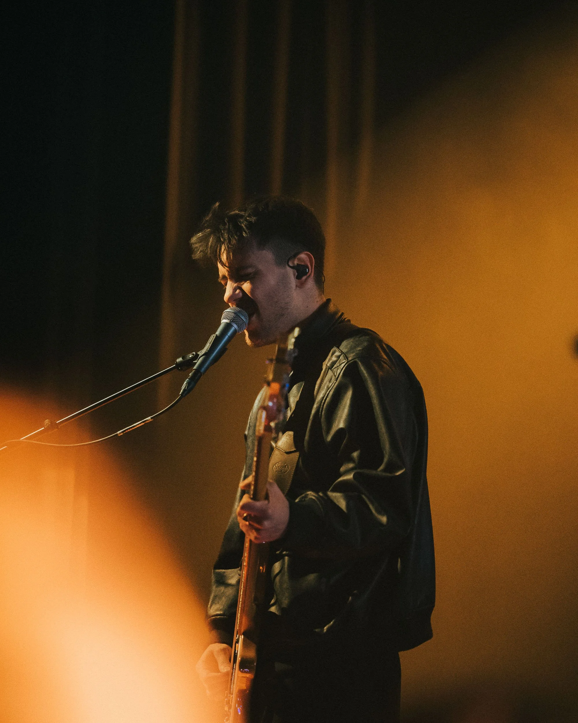 Musician singing into a microphone while playing an electric guitar on stage with warm lighting.