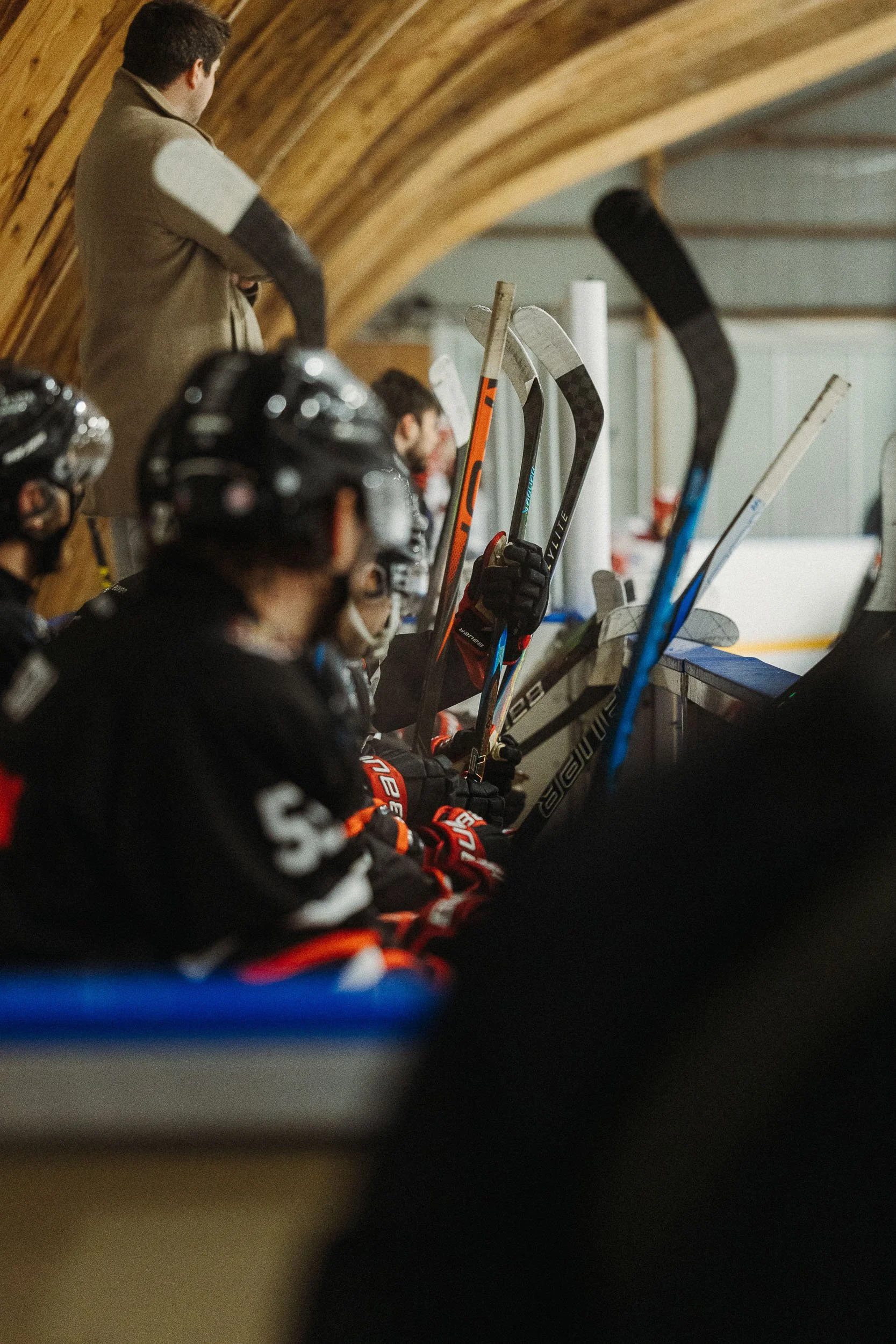 Ice hockey players on the bench with hockey sticks, wearing helmets and gear, watching a game on the ice, with coach standing nearby.