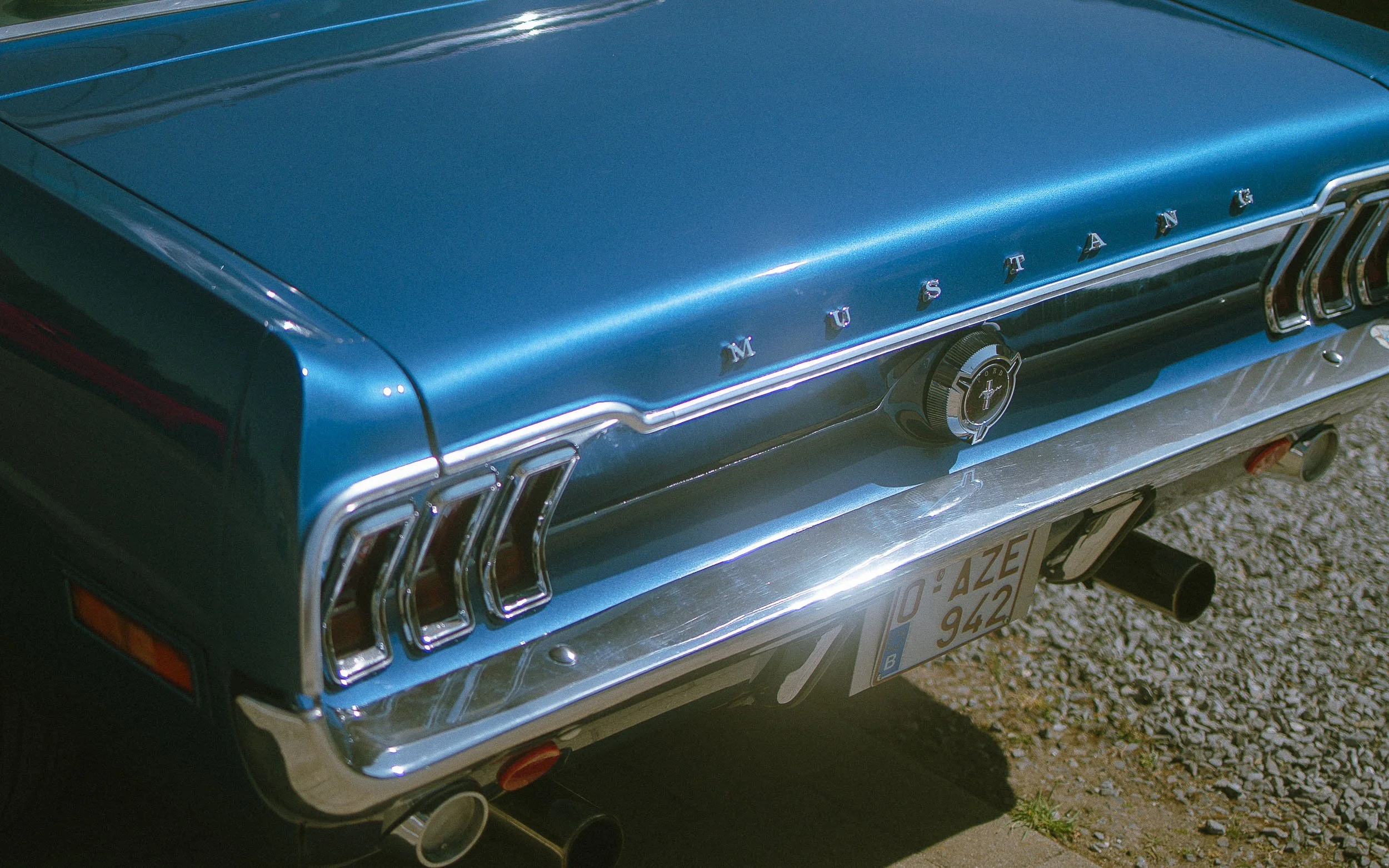 Back of a vintage blue Ford Mustang with chrome details, taillights, a license plate, and dual exhaust pipes.