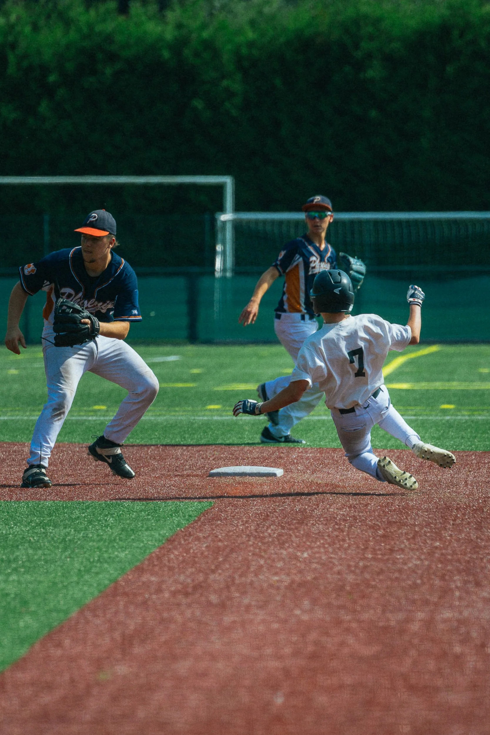 A baseball player in a white uniform sliding into a base while a player in a navy uniform prepares to receive the ball. Two other players are in the background on a baseball field.