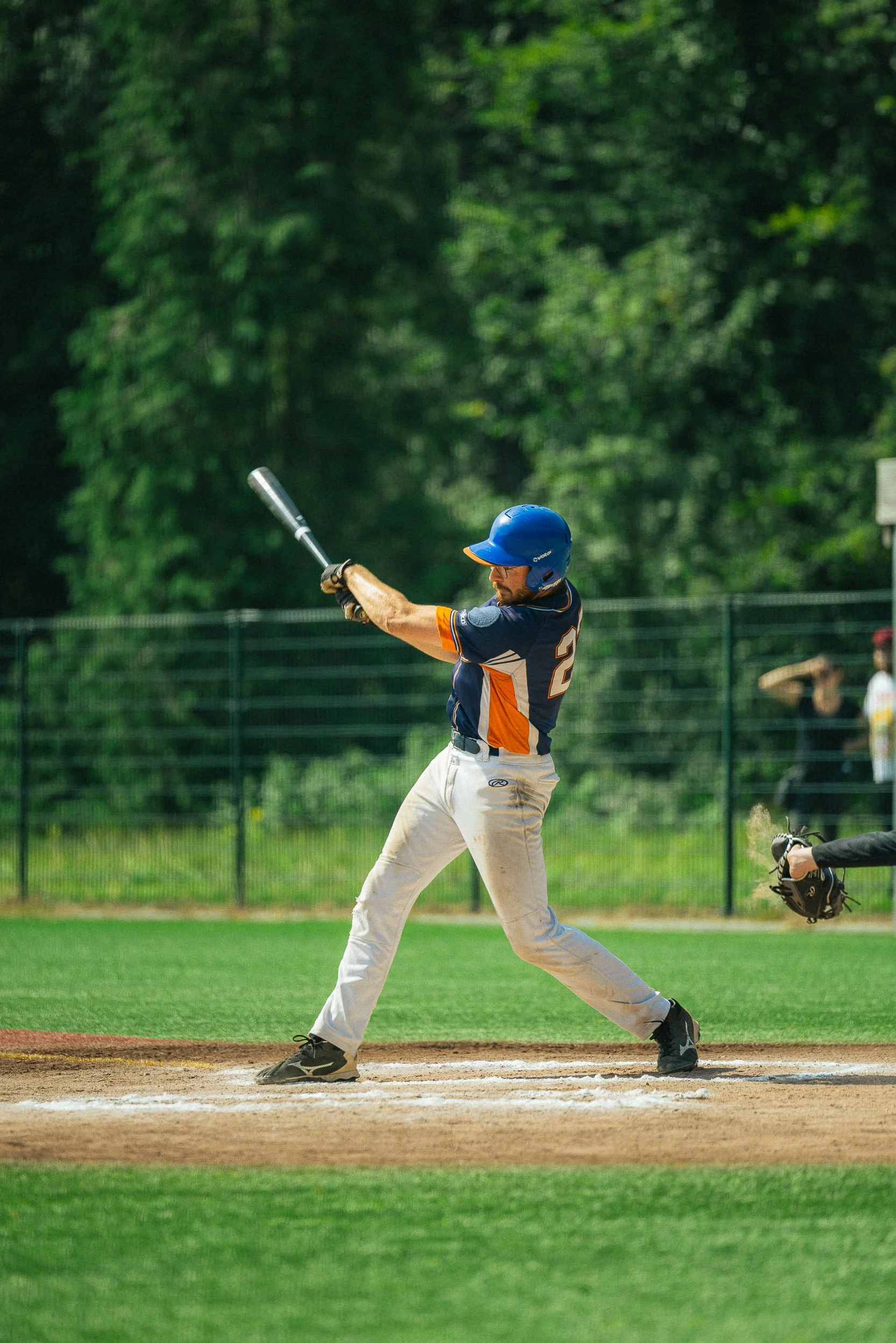 A male baseball player in a blue and orange uniform, wearing a helmet, swings a bat at a pitch on the field with a green outfield and trees in the background.