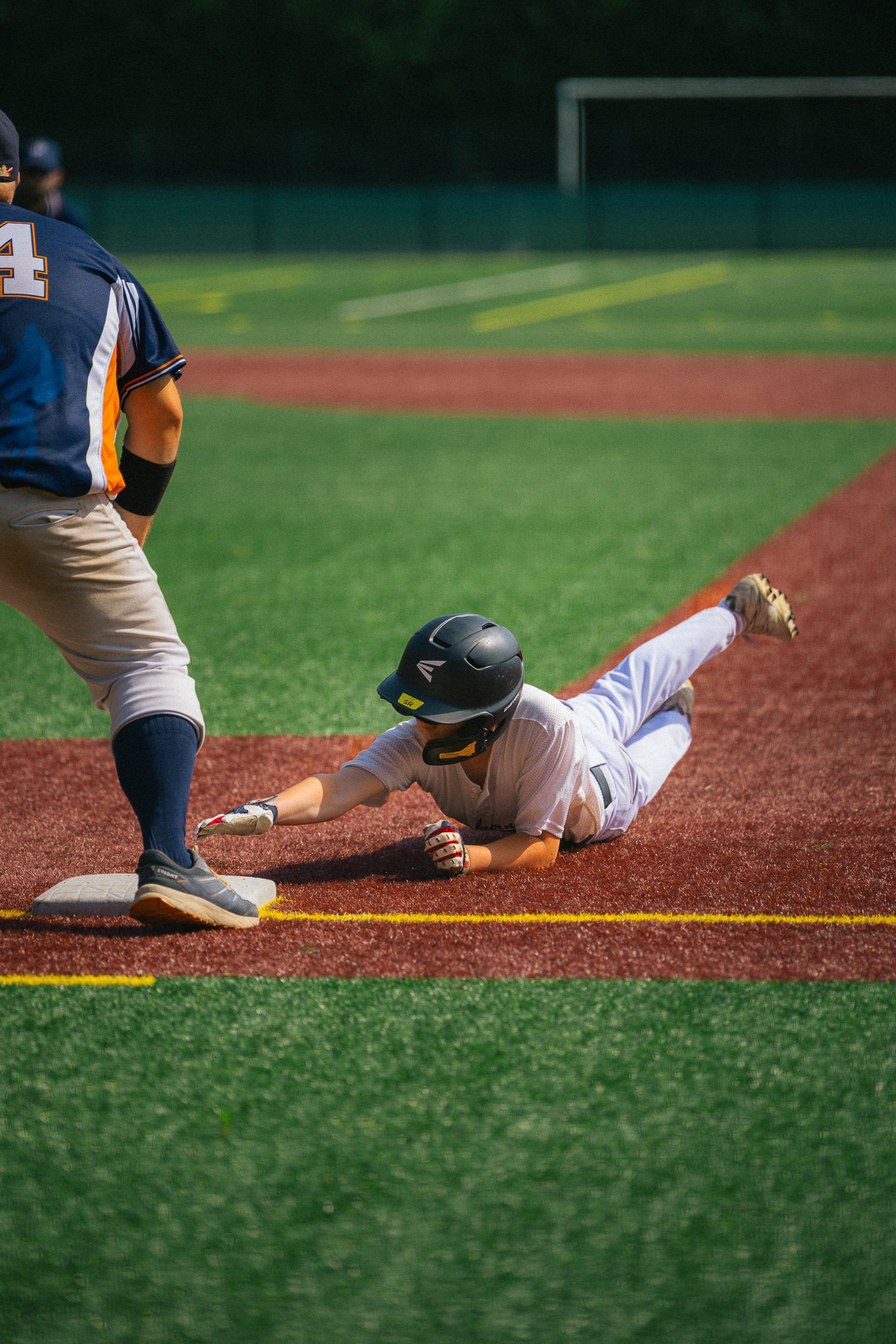 A baseball player wearing a white uniform and black helmet is sliding into a base on a baseball field, while another player in a navy jersey and beige pants is standing near the base.