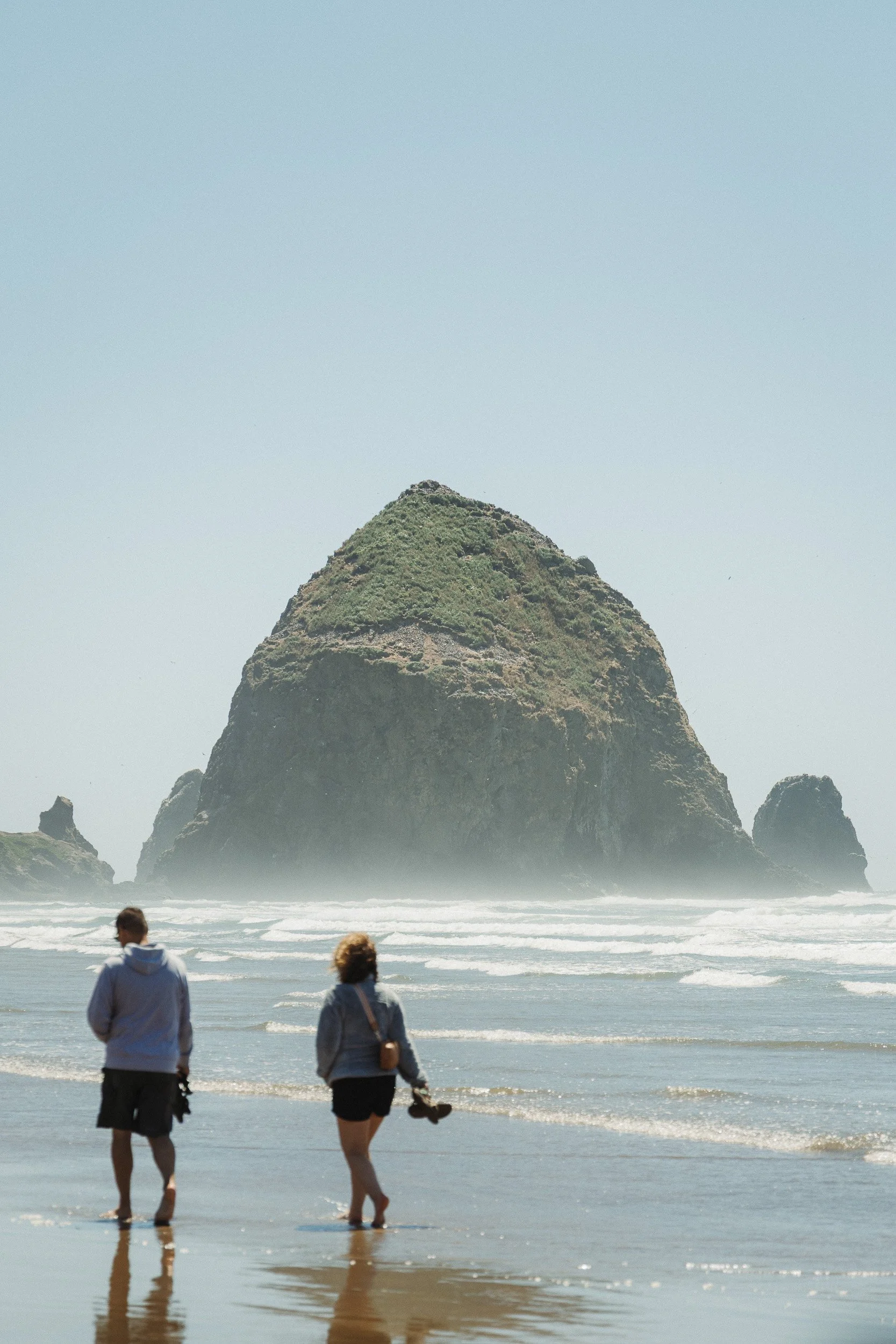 Two people walking along the shoreline with a large rock formation in the ocean behind them.