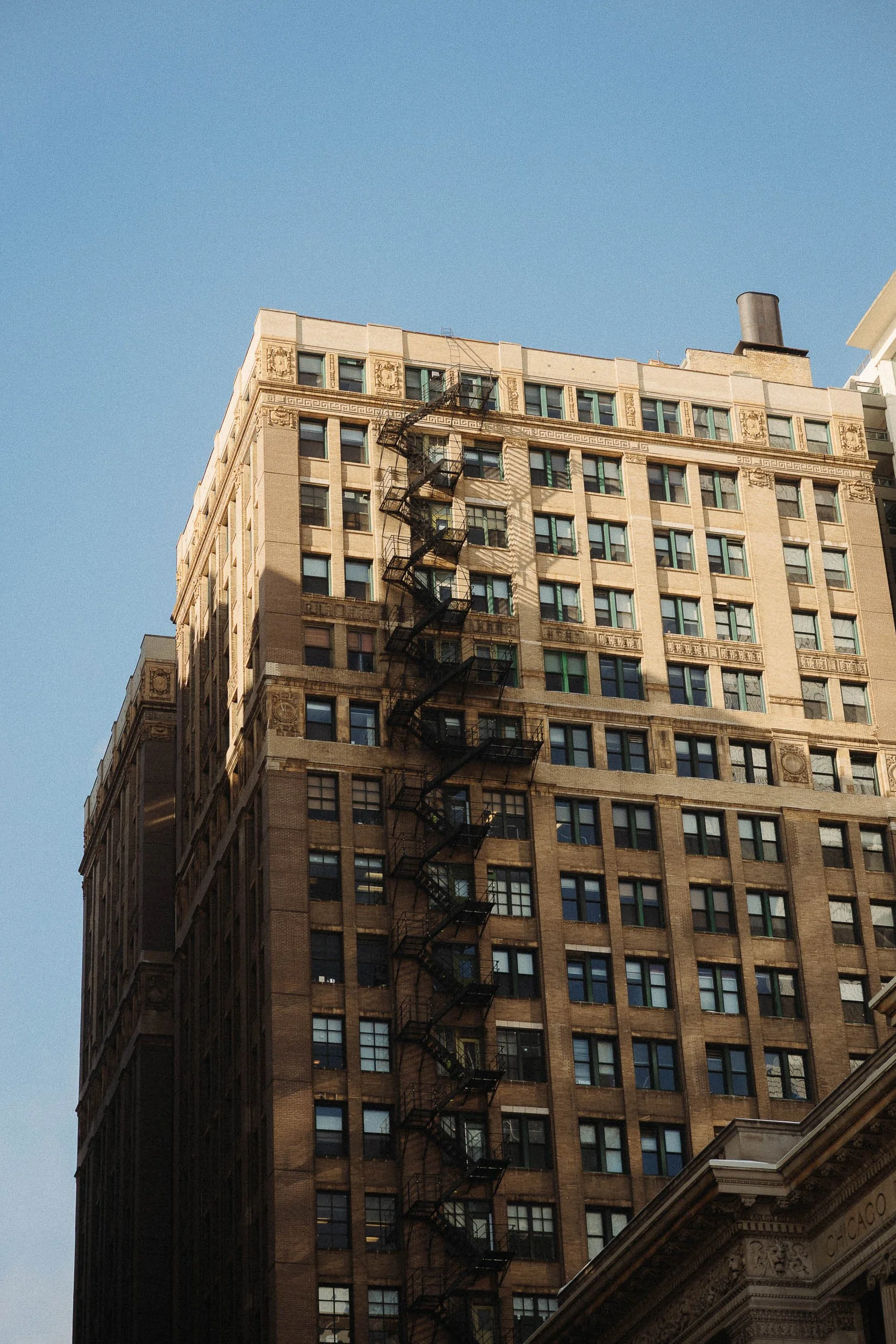 A tall, historic building with beige and brown brick facade, multiple windows, decorative architectural details, and a fire escape running vertically down the front. Part of a building with ornate molding at the bottom right corner.