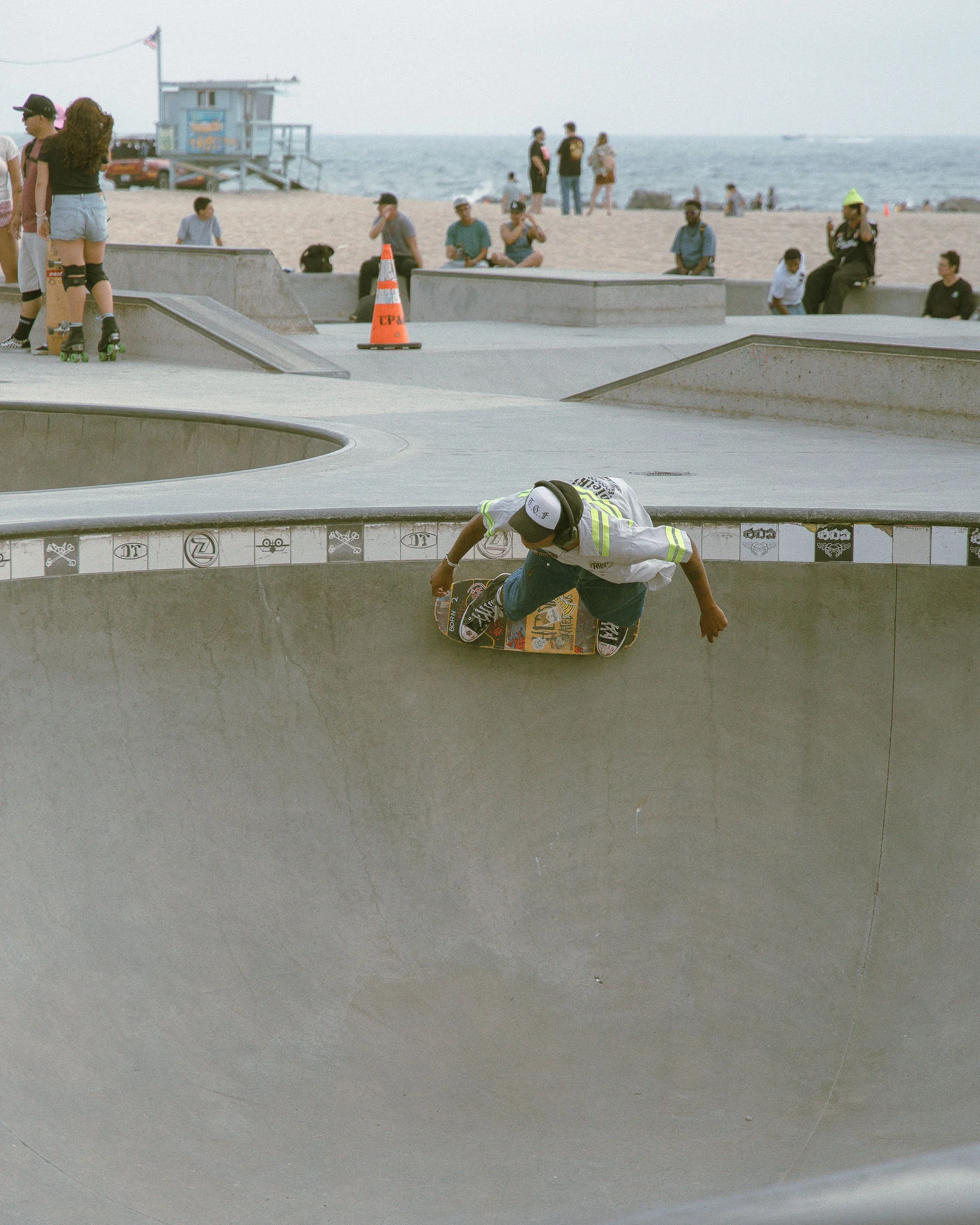 A skateboarder performs a trick in a skate park near the beach, with people sitting and walking in the background.
