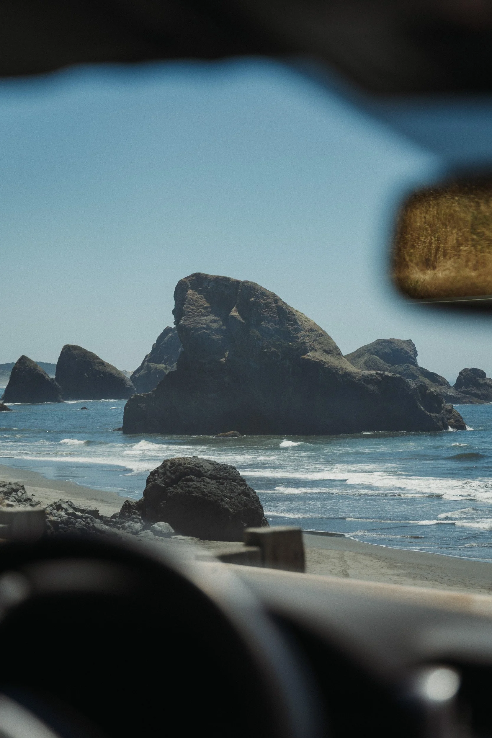 View of rocky ocean coastline seen from inside a vehicle, with rocks in the water and sandy beach in the foreground.