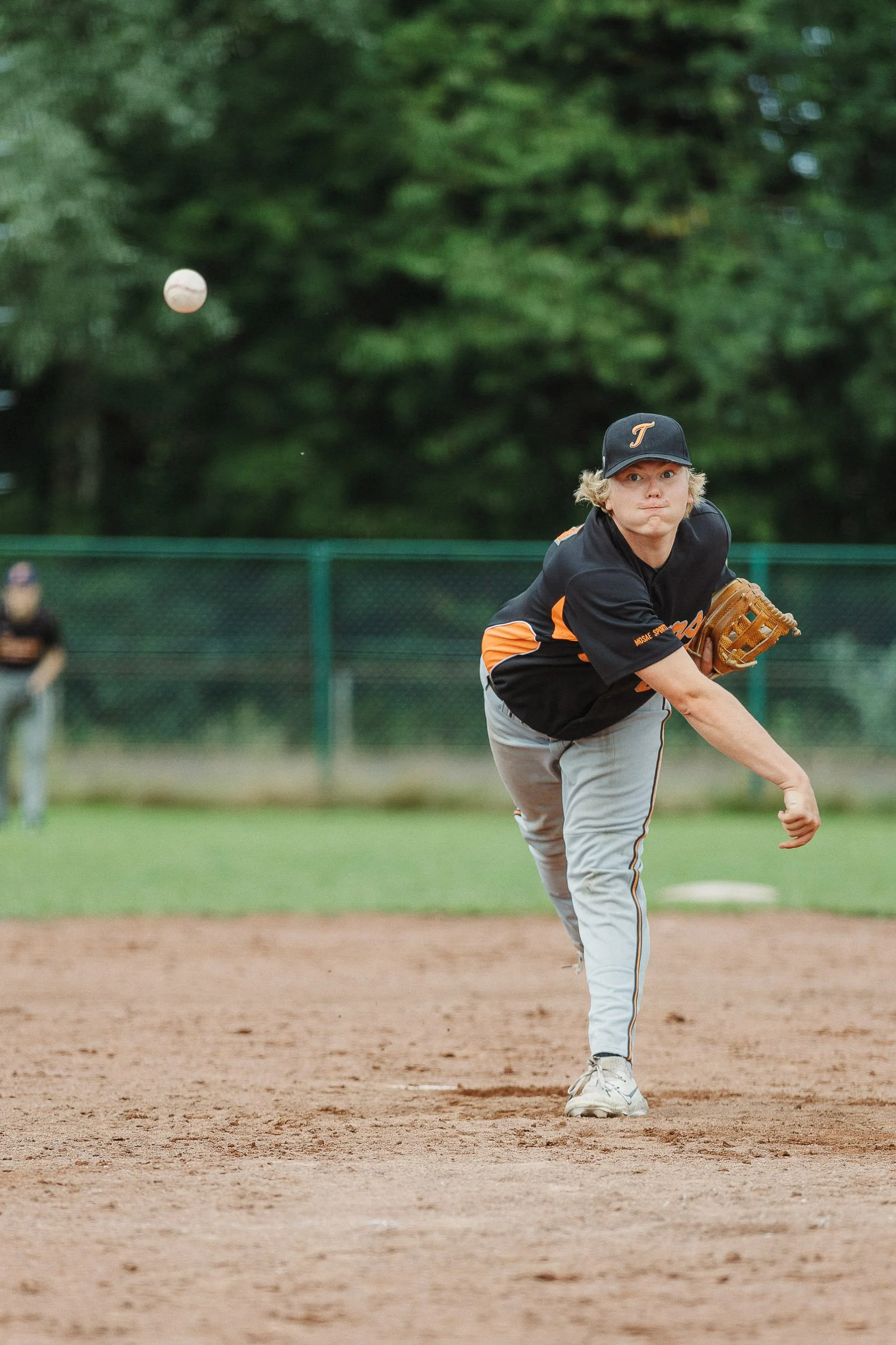 A baseball player in a black and orange uniform pitching on a baseball field with green trees in the background.