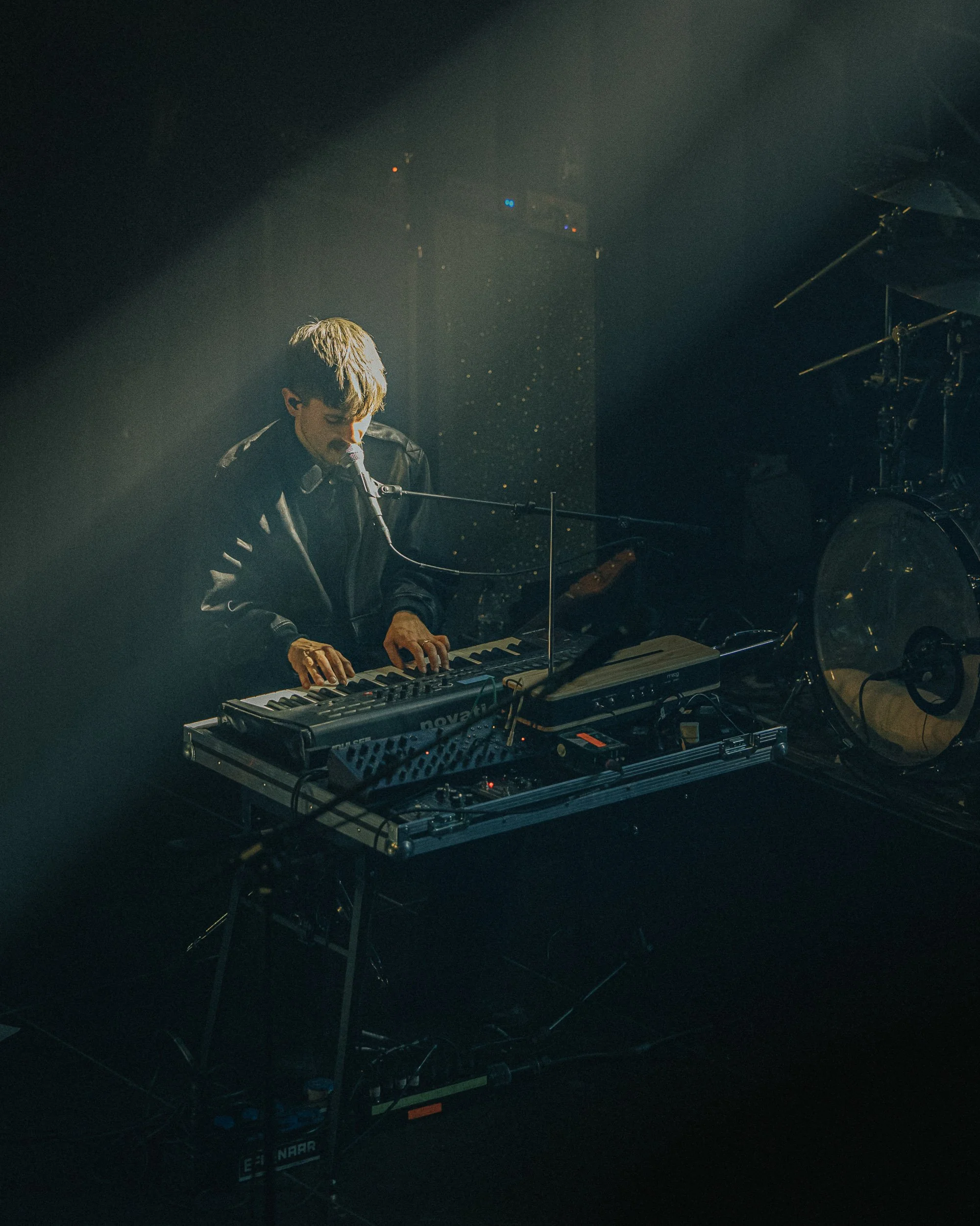 A musician playing keyboard on stage with a microphone, lit by soft spotlight, with musical equipment around.