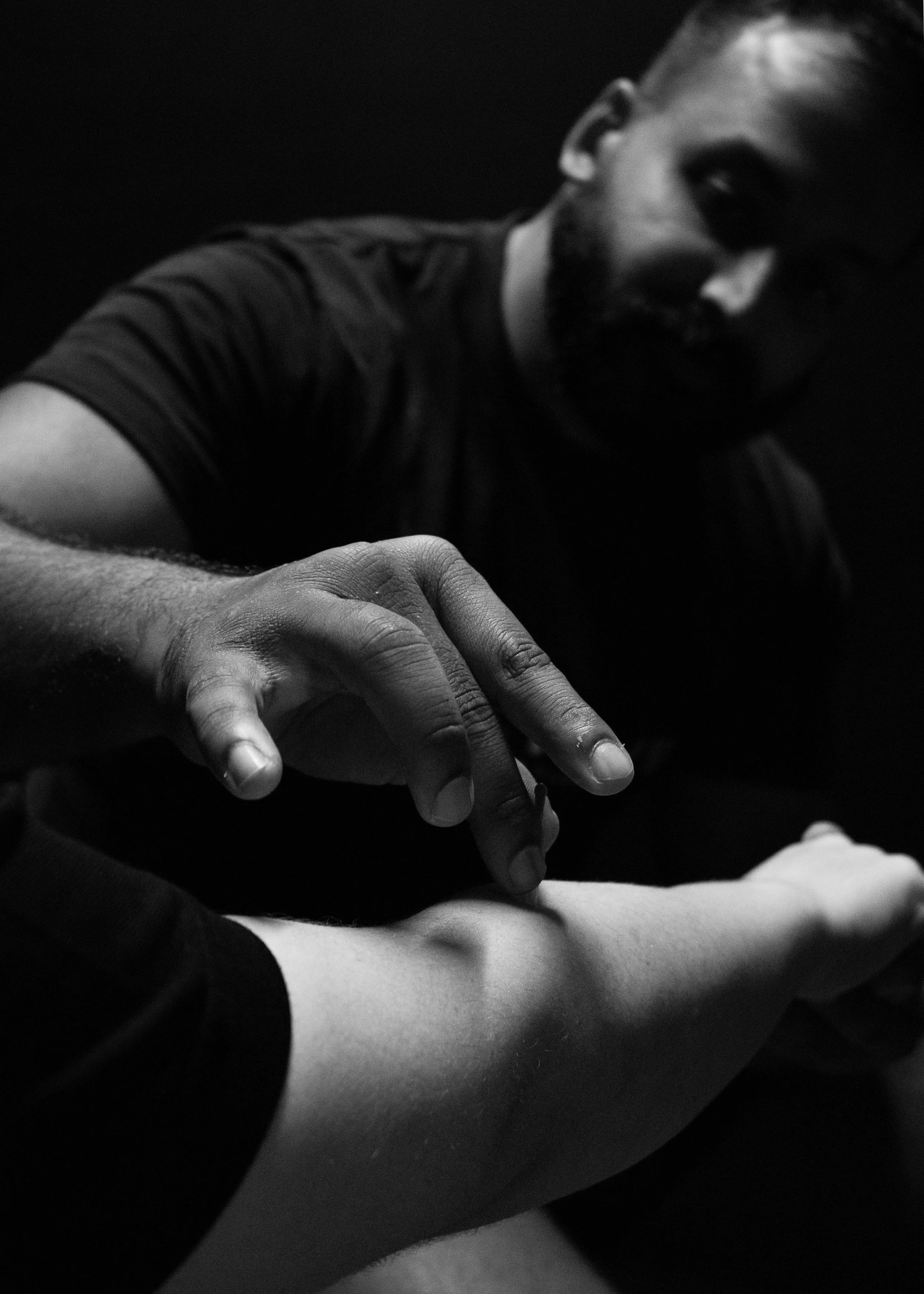 Close-up black and white photo of a man with facial hair reaching out his hand toward the camera, partially obscured face, dark clothing, focusing on his hand and arm.