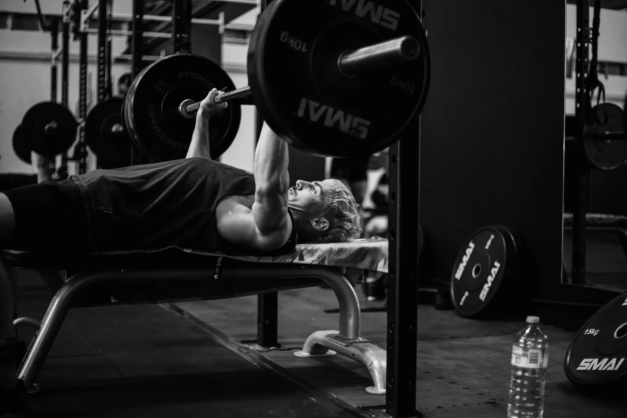 A person lying on a workout bench lifting a barbell with weights in a gym. The person is smiling, and there are weight plates next to the bench, as well as a water bottle on the ground.