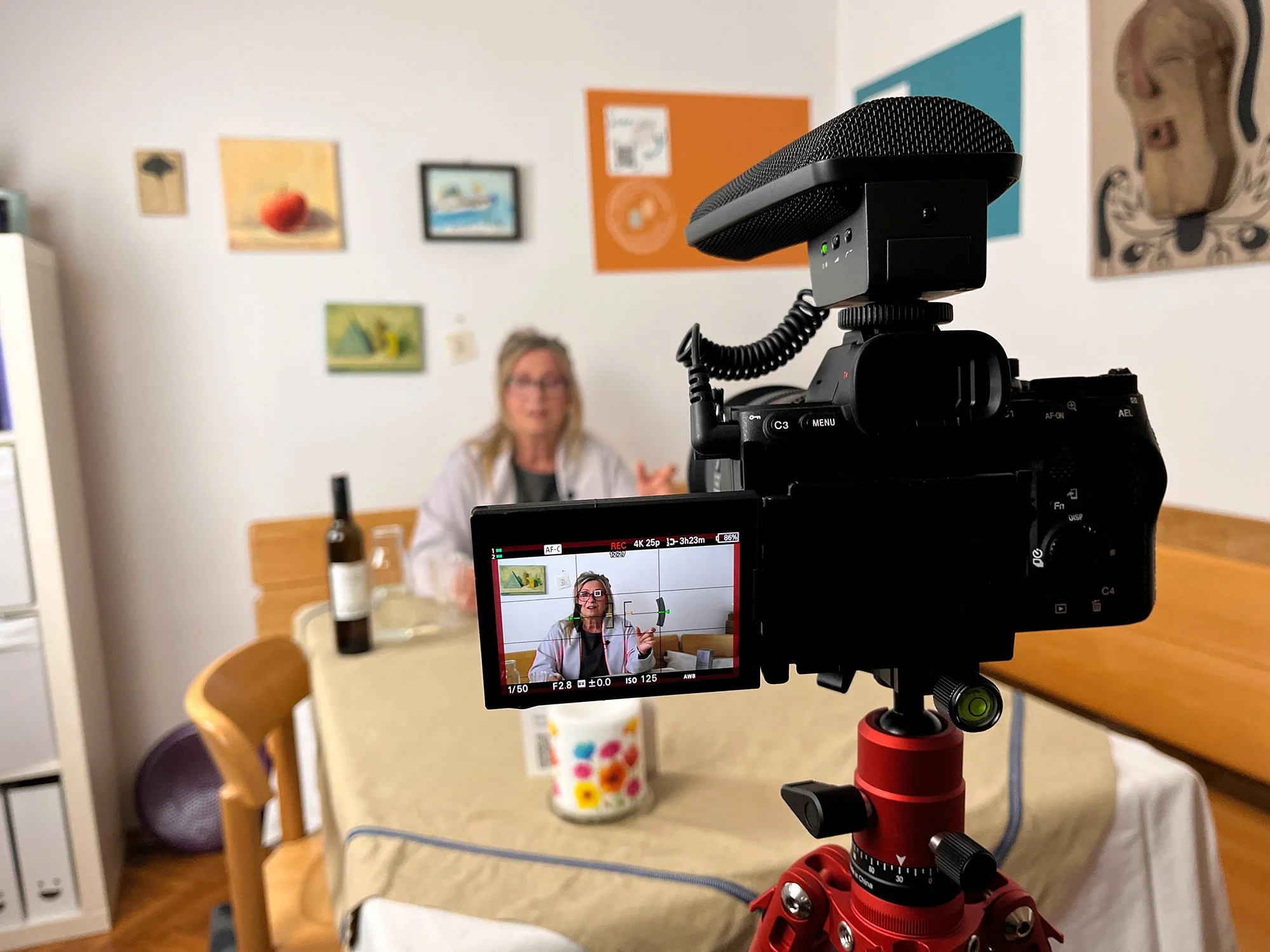 Eine Kamera auf einem Stativ filmt eine Frau, die an einem Tisch sitzt. Auf dem Tisch stehen eine Weinflasche und ein Glas. Die Wand im Hintergrund ist mit Bildern dekoriert. Auf dem Kameradisplay sieht man die Frau im Fokus.