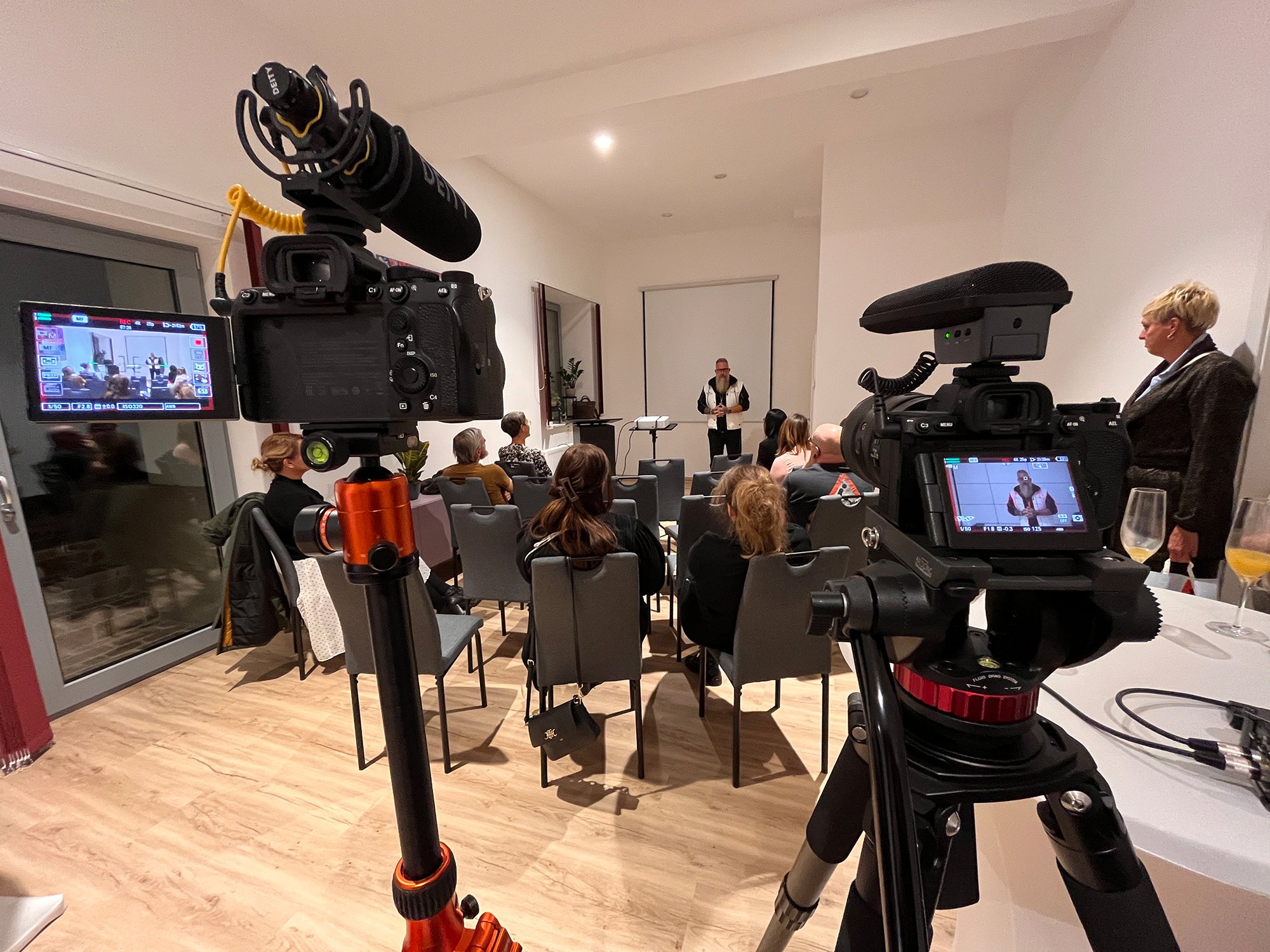 Ein Raum mit mehreren Personen, die einer Präsentation oder Veranstaltung folgen. Zwei Kameras auf Stativen filmen die Szene. Eine Person steht vorne und spricht zum Publikum. Einige Frauen sitzen, und eine Frau steht an der Seite mit Getränkegläsern