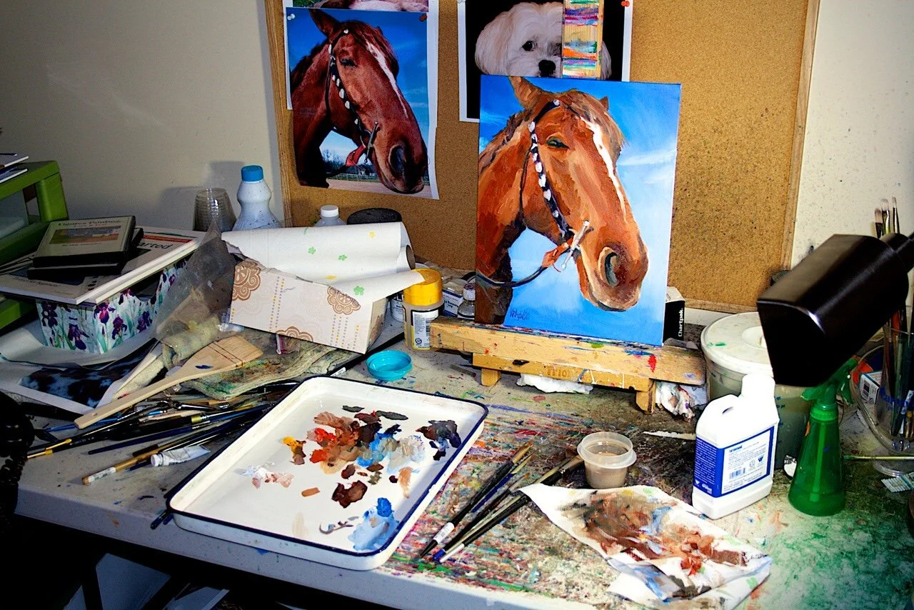 Art studio desk with a painting of a brown horse on a small easel, paint supplies, brushes, and reference photos of the horse on the wall.