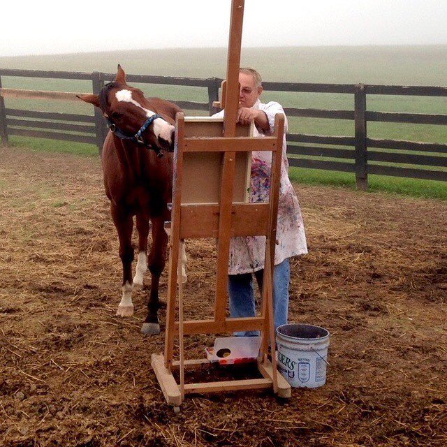 Ron Krajewski painting outdoors near a wooden fence with Metro the painting racehorse beside him