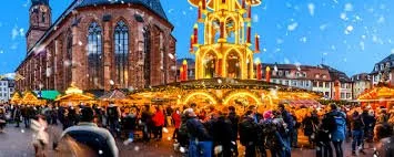 A Christmas market scene with a decorated carousel, illuminated stalls, and a crowd of people enjoying the festive atmosphere outdoors during snowfall.