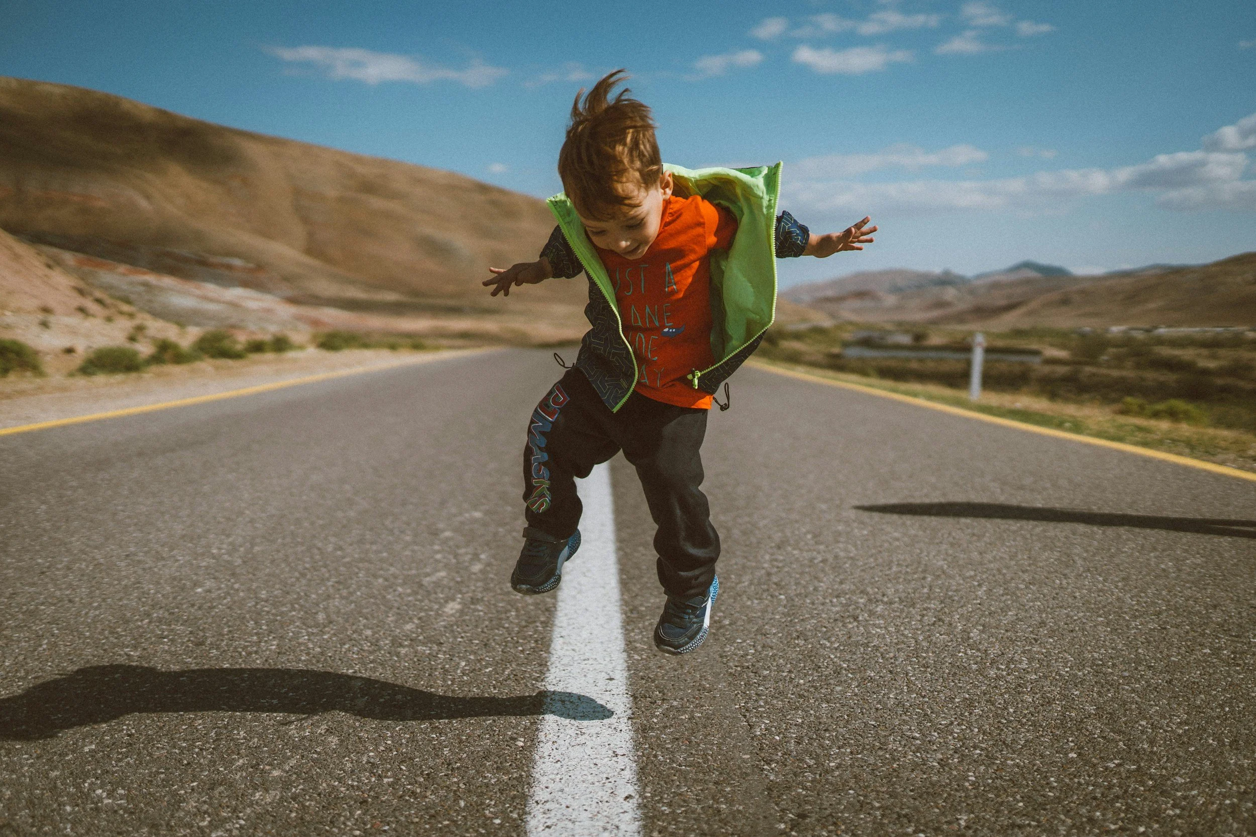 A young boy jumping in the middle of a deserted road in a scenic desert landscape with hills in the background.