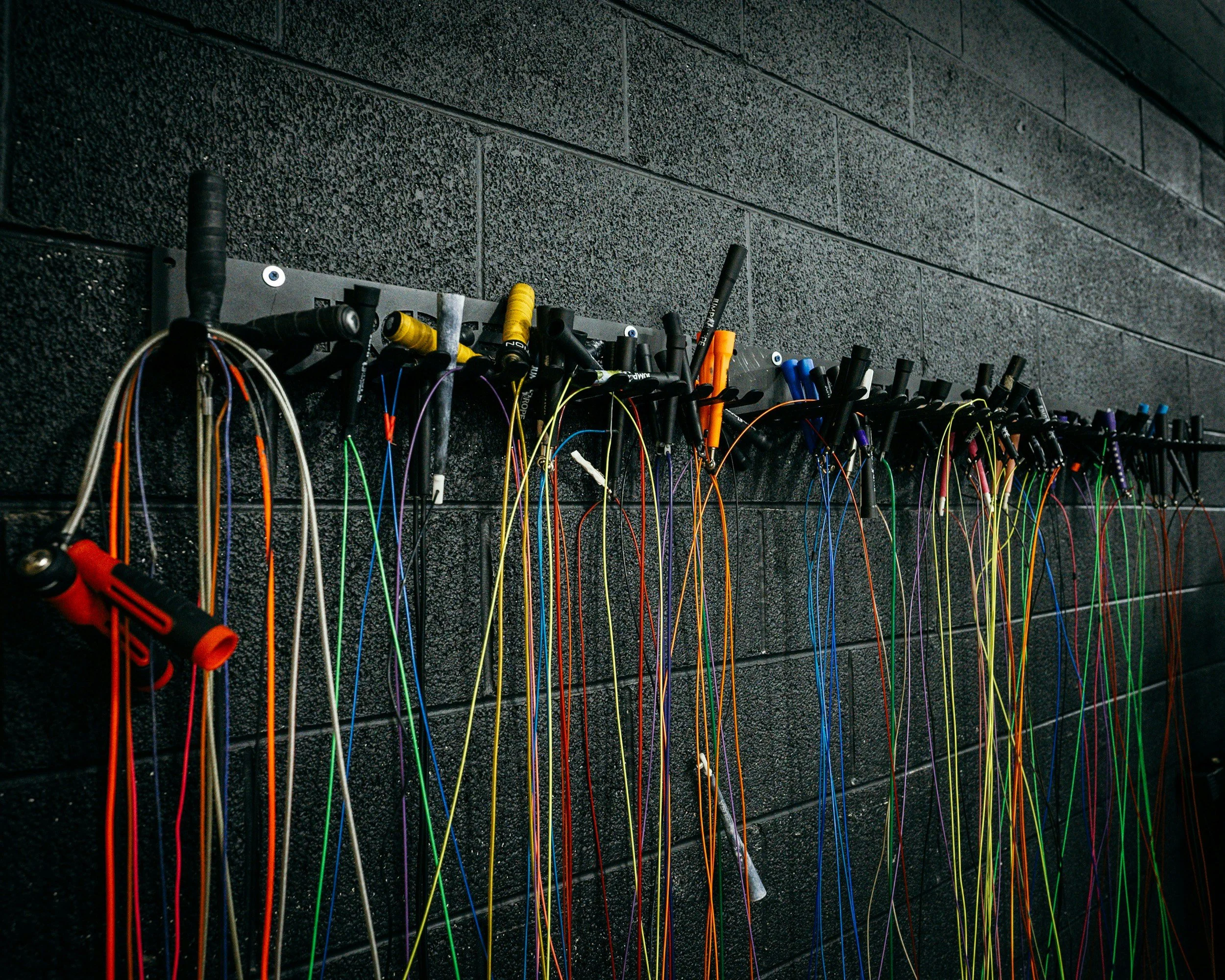 A wall-mounted rack holding various colorful bungee cords and handles, with a black textured wall in the background.