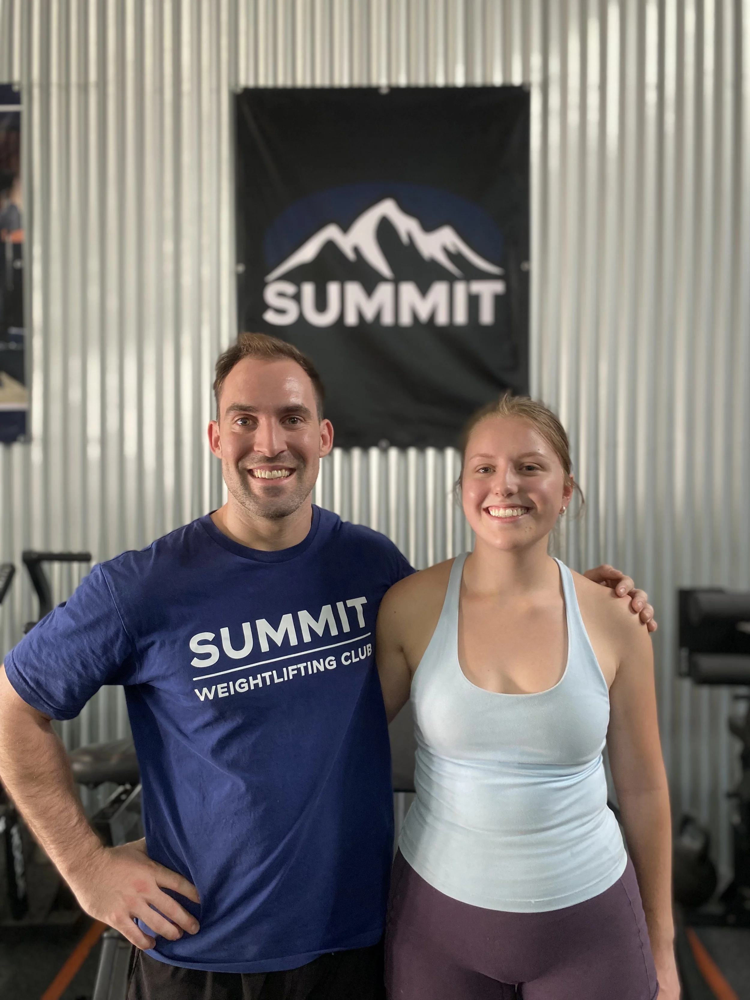 A man and woman smiling at the camera in a gym with a 'SUMMIT' banner behind them, wearing workout clothes.